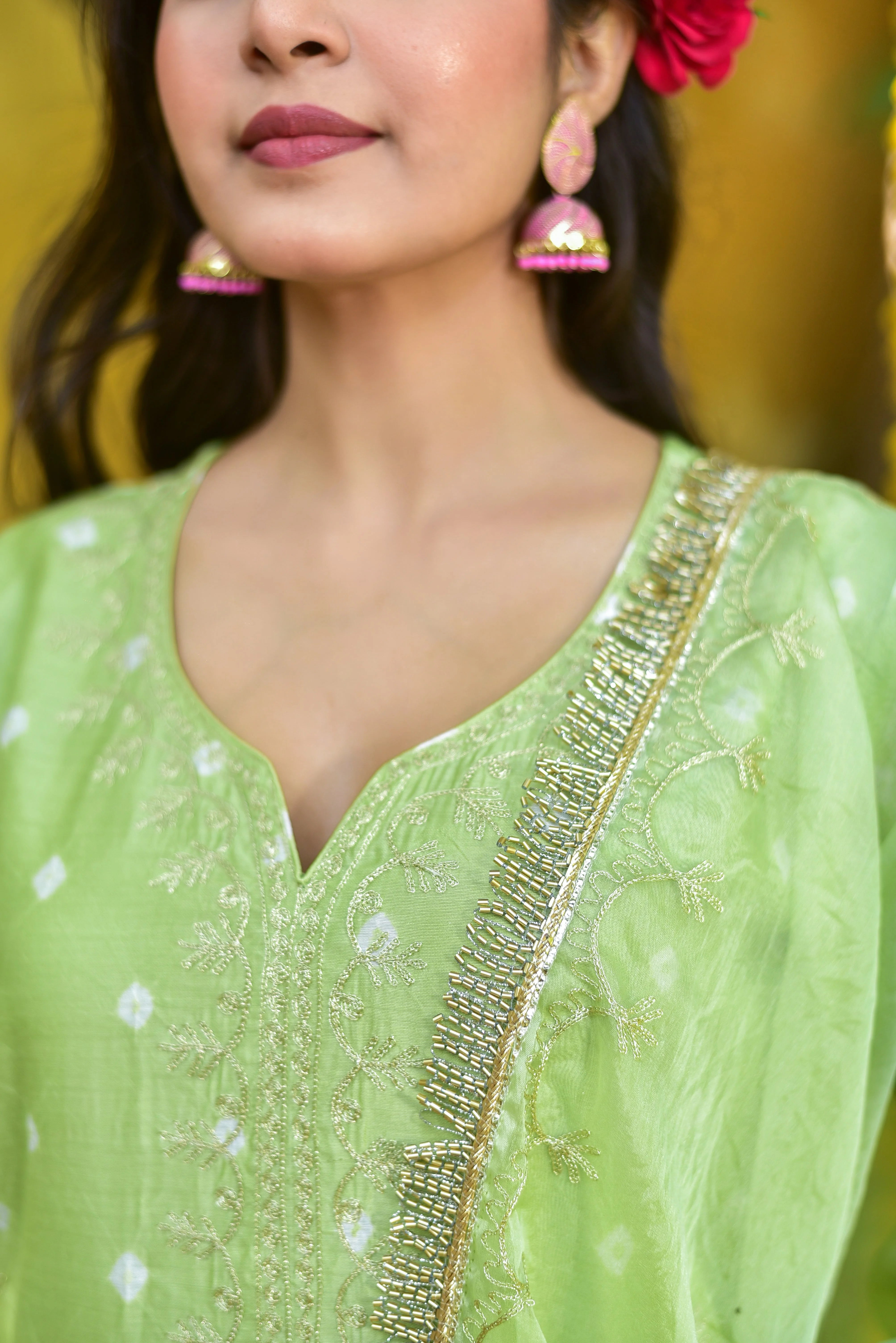 This is a high-quality image of a woman wearing a green bandhej a-line kurta set, showcasing the intricate details of the traditional Indian garment. The image is well-lit, with the woman standing in front of a plain background that allows the vibrant colors of the kurta to take center stage.
The woman's long, dark hair cascades down her back, complemented by a delicate pink flower tucked behind her ear. Her makeup is subtle yet elegant, with a focus on enhancing her natural features.