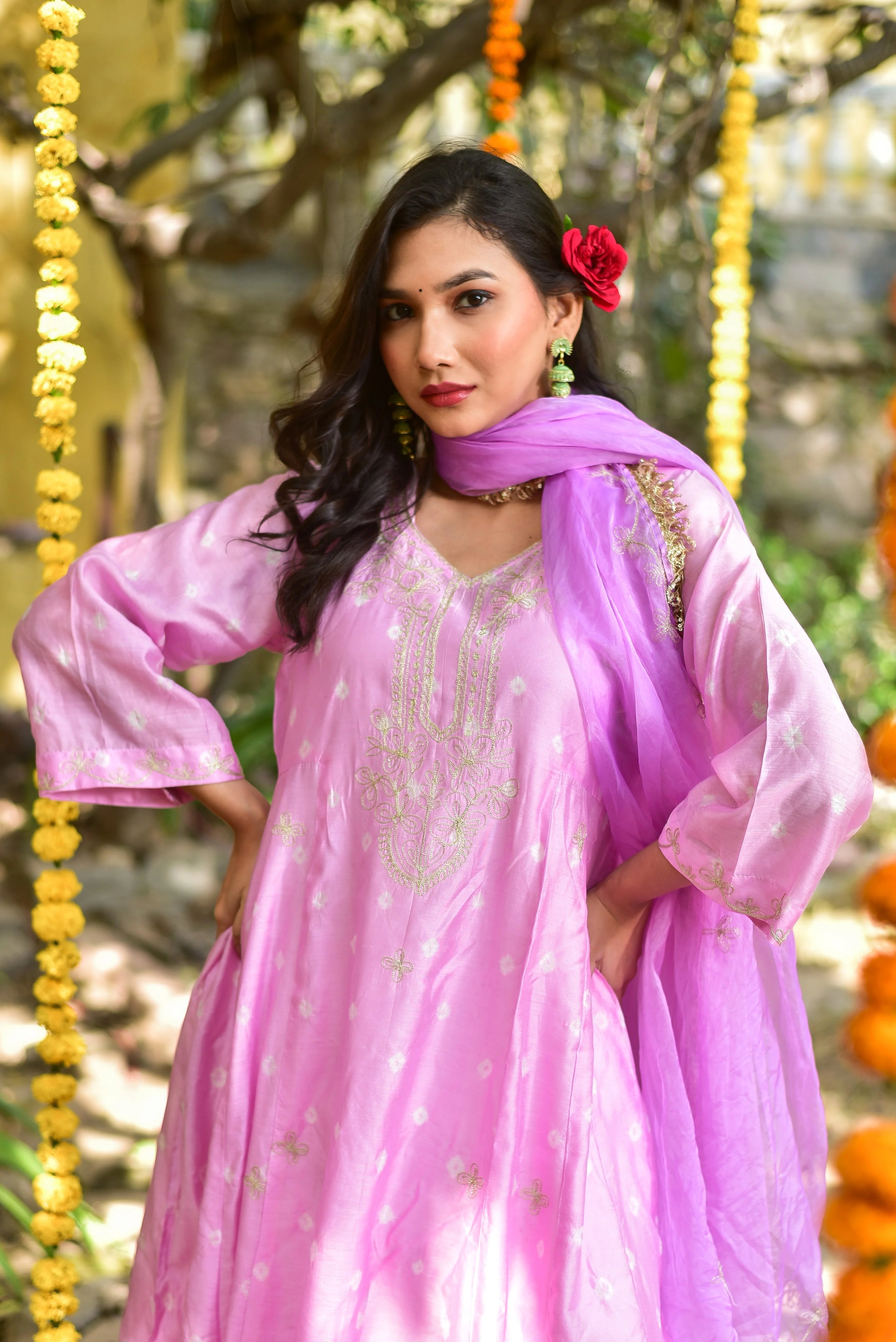 The image features a woman wearing a stunning pink salwar kameez, a traditional Indian outfit consisting of a long tunic (kameez) and matching pants (salwar). The outfit is made of silk or organza fabric, which drapes elegantly on the woman's body.
The kameez boasts a V-neckline and three-quarter sleeves, with intricate Mukesh work adorning the chest, neckline, and sleeves. The Mukesh work is a traditional Indian embroidery technique that uses