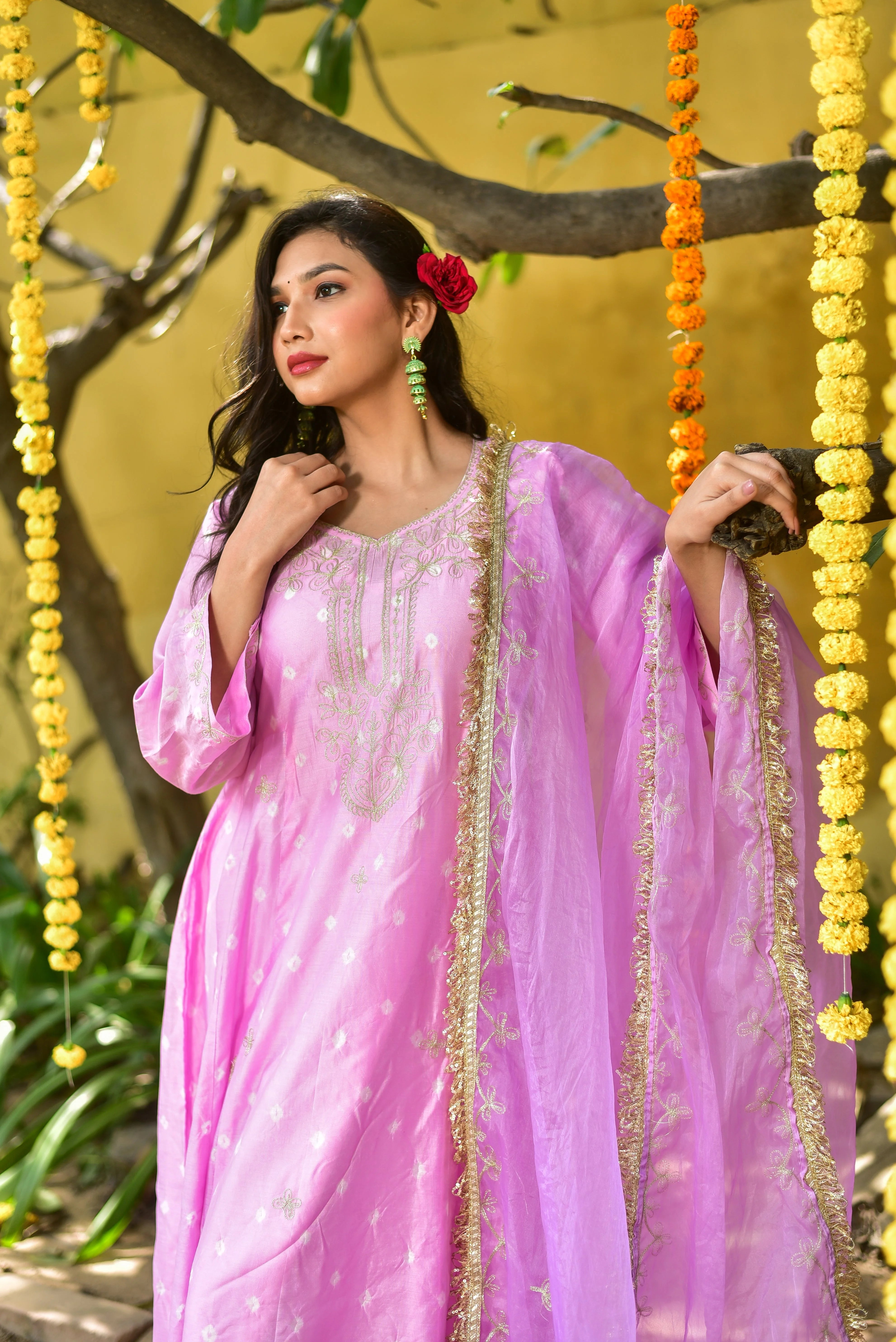 The image features a woman dressed in a stunning pink salwar kameez, exuding elegance and sophistication. The long, flowing dress boasts intricate Mukesh work, a traditional Indian embroidery technique that adds a touch of luxury to the garment. The V-neckline and three-quarter sleeves create a flattering silhouette, while the matching dupatta with golden embroidery and a delicate border adds a pop of color and texture to the overall ensemble.
The model's dark hair is adorned with a red rose, adding a subt