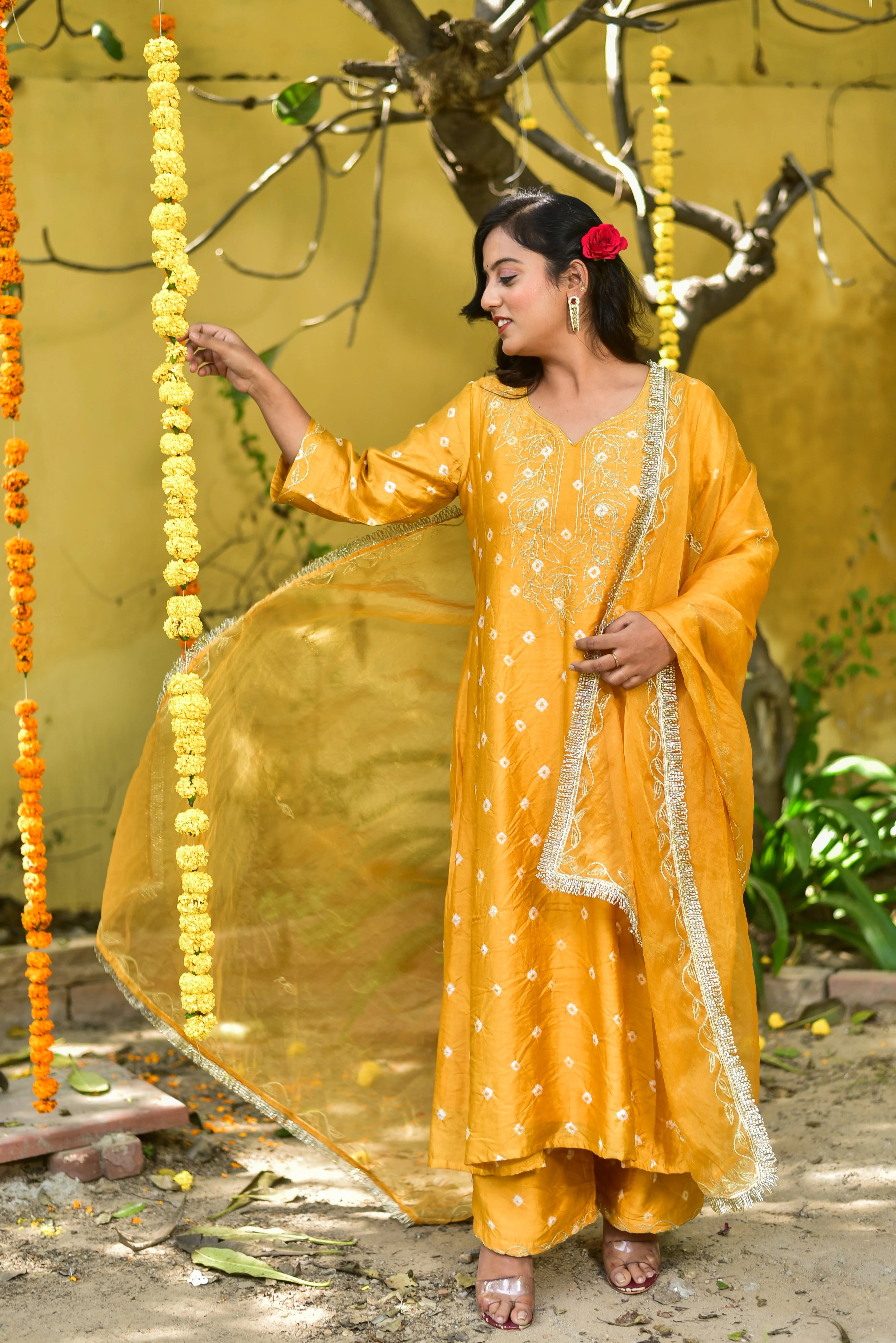 The image depicts a woman in a traditional Indian outfit, showcasing the Mustard Yellow Bhandej Kurta Set. The set features a long-sleeved kurta with a V-neckline and a matching pair of pants, both adorned with intricate white bandhani designs. The outfit is complemented by a sheer yellow dupatta with a delicate fringe trim, adding a touch of elegance to the overall look.
The woman's dark hair is styled with a red flower tucked behind her ear, and she accessorizes with silver earrings and clear heels.