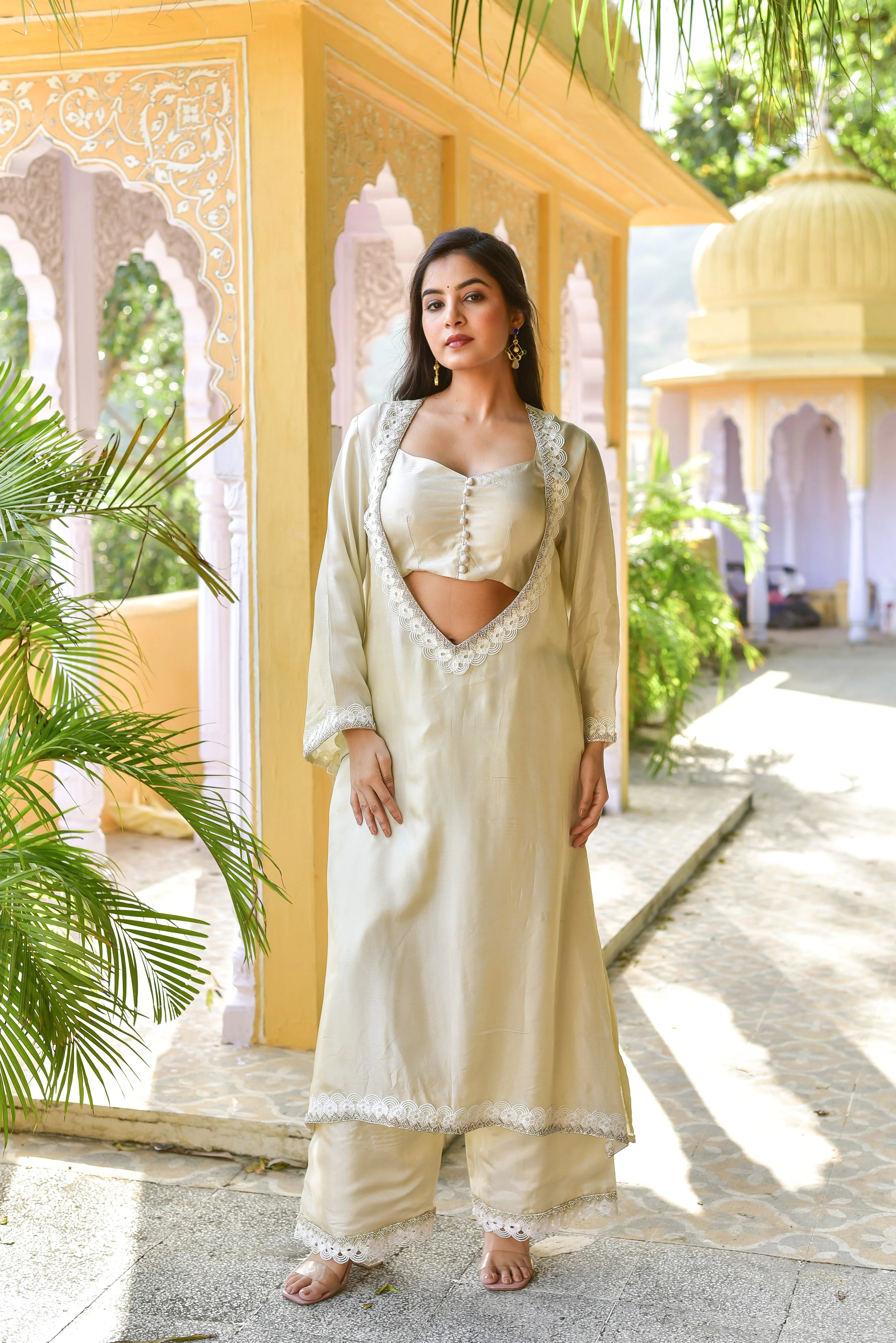Woman in a traditional outfit standing in a decorative outdoor setting with plants and architecture.