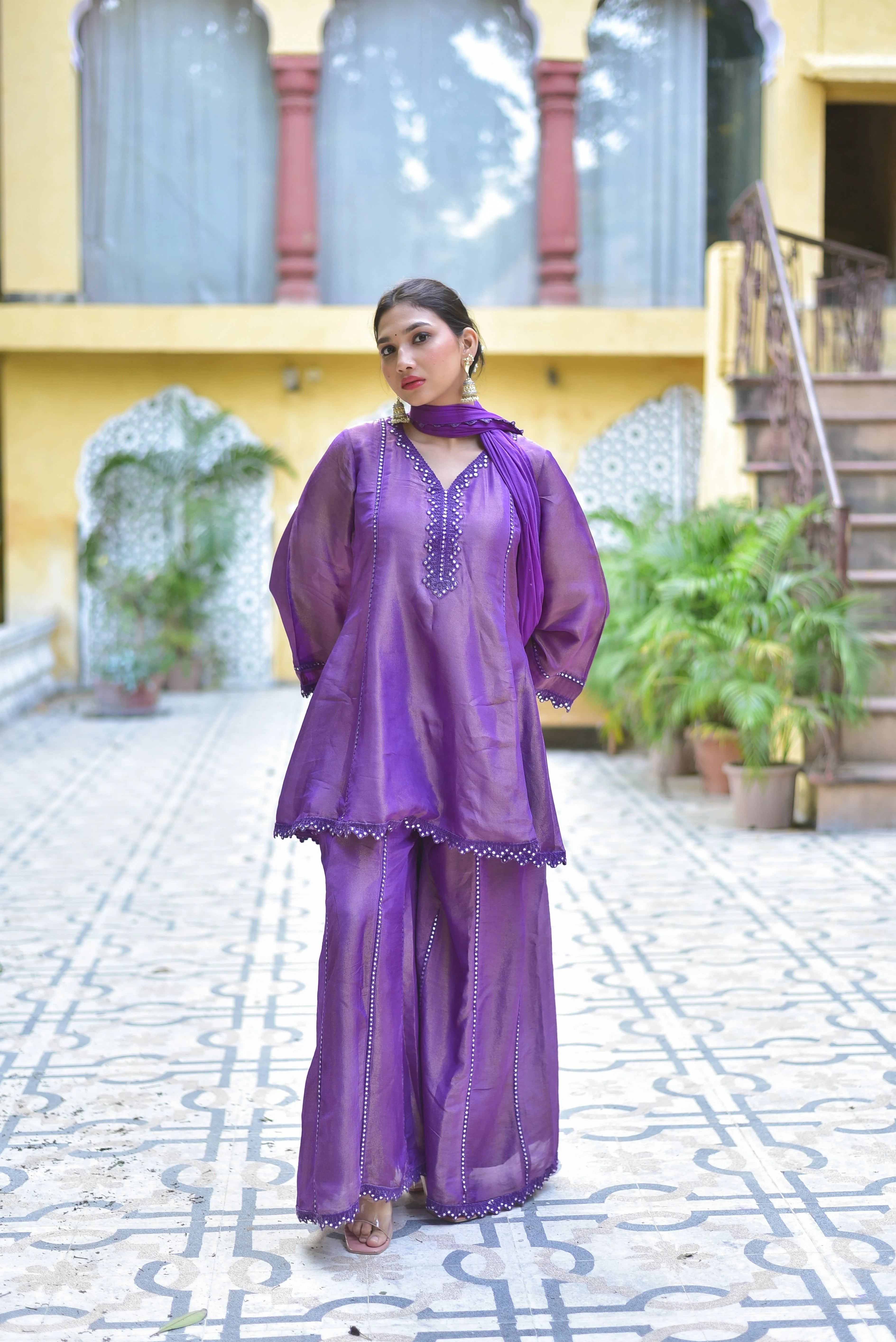 The image showcases a woman standing in front of a building, wearing a stunning purple zari silk kurta sharara set. The outfit features a V-neckline with intricate embroidery, long sleeves, and a flowing sharara that falls just above the ankles. The set is crafted from luxurious zari silk fabric, exuding elegance and sophistication.
In the background, a yellow building with arched windows and a staircase leading to an open doorway provides a warm and inviting setting.