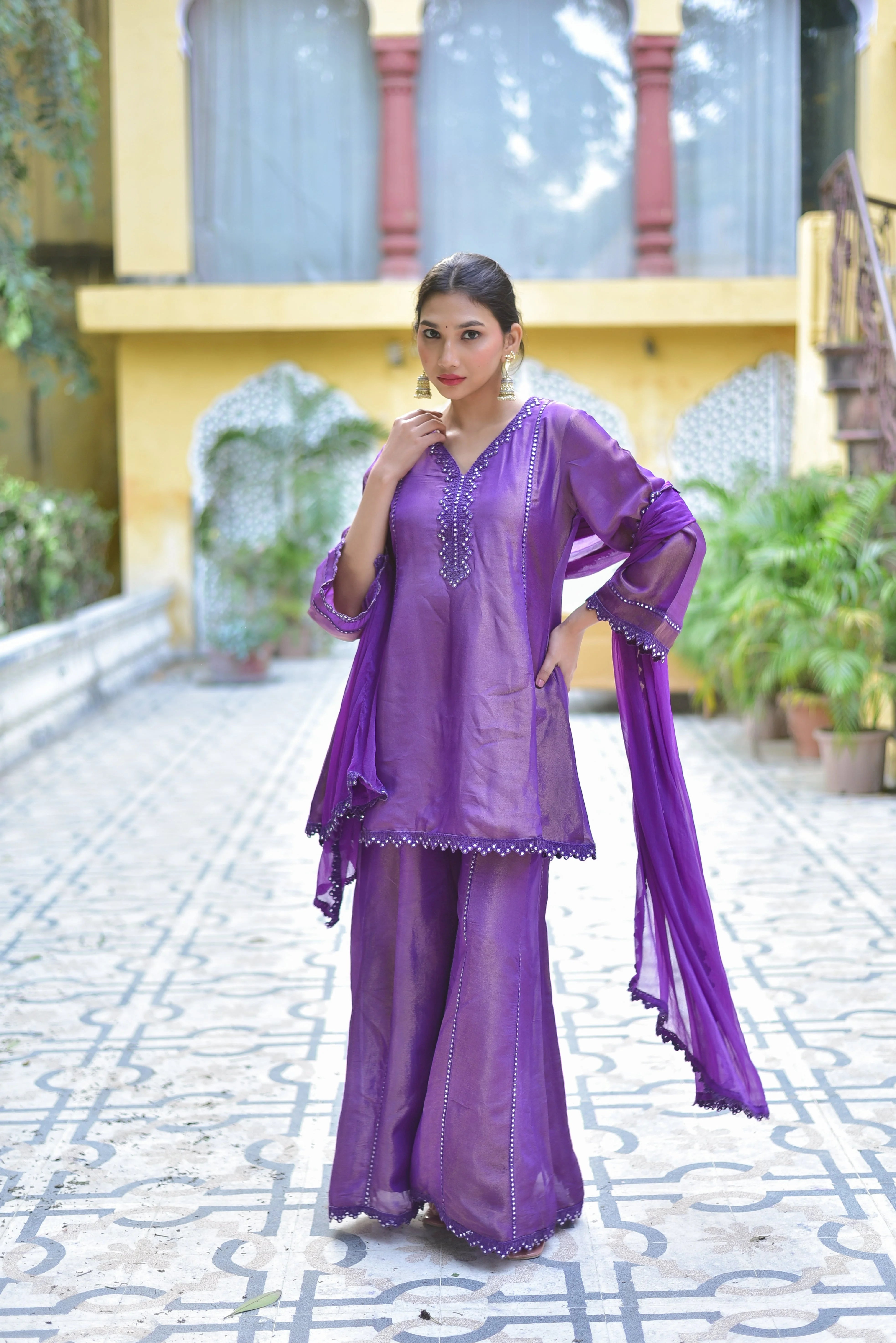 The image features a woman in a stunning purple Zari Silk Kurta Sharara Set, showcasing traditional craftsmanship and contemporary elegance. The set consists of a kurta with a V-neckline and long sleeves, adorned with intricate zari embroidery on the yoke and hem. The matching sharara pants have a flared silhouette, while the dupatta is draped elegantly over the shoulders, adding a touch of sophistication to the overall look.