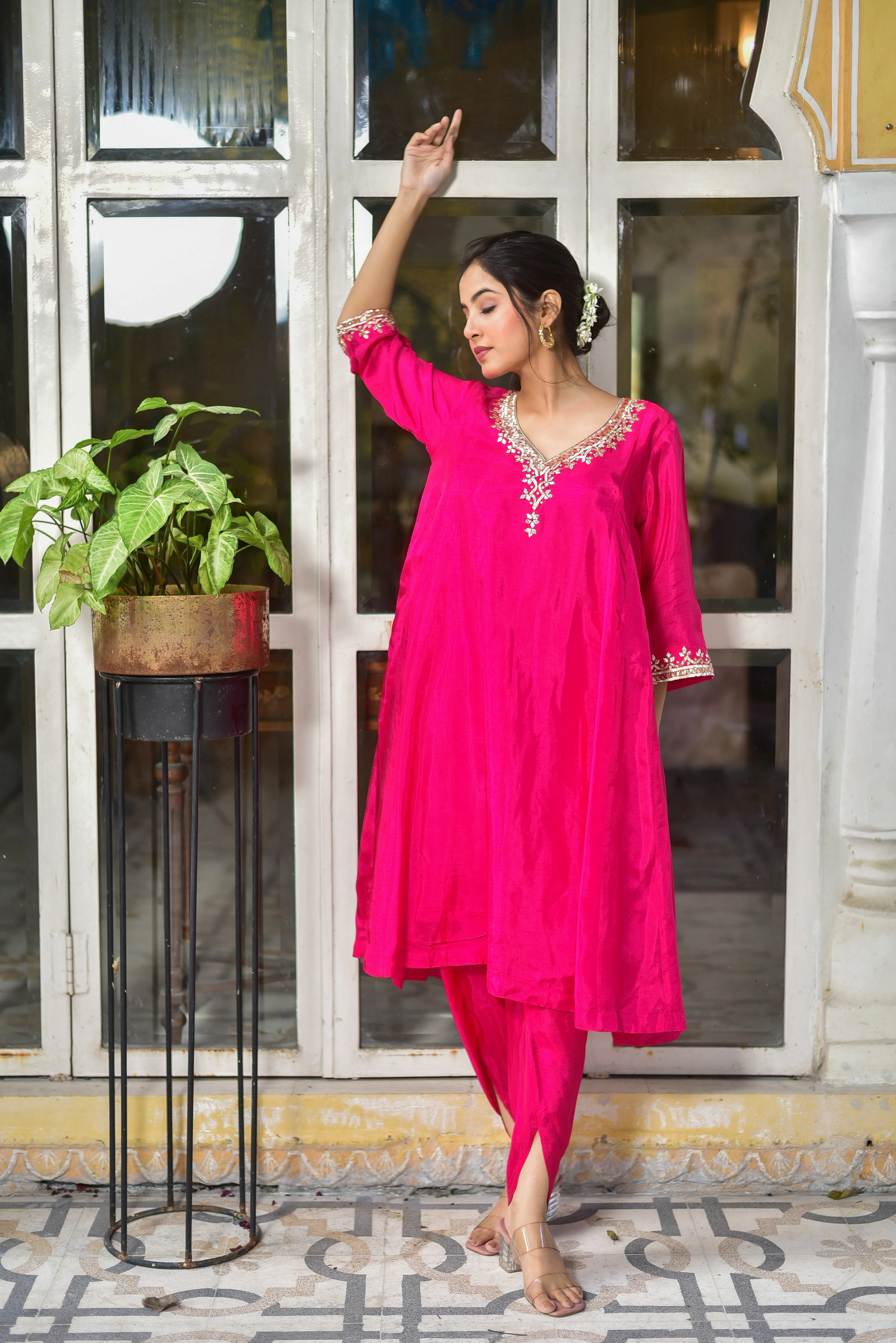 The image depicts a woman standing in front of a window, wearing a stunning Rani Pink Punjabi Style Kurta & Dhoti Set. The set is crafted from luxurious silk, featuring a V-neckline adorned with intricate white embroidery that adds a touch of elegance. The kurta boasts a loose, flowing fit with three-quarter sleeves, while the dhoti pants are designed with a stylish slit at the ankle.