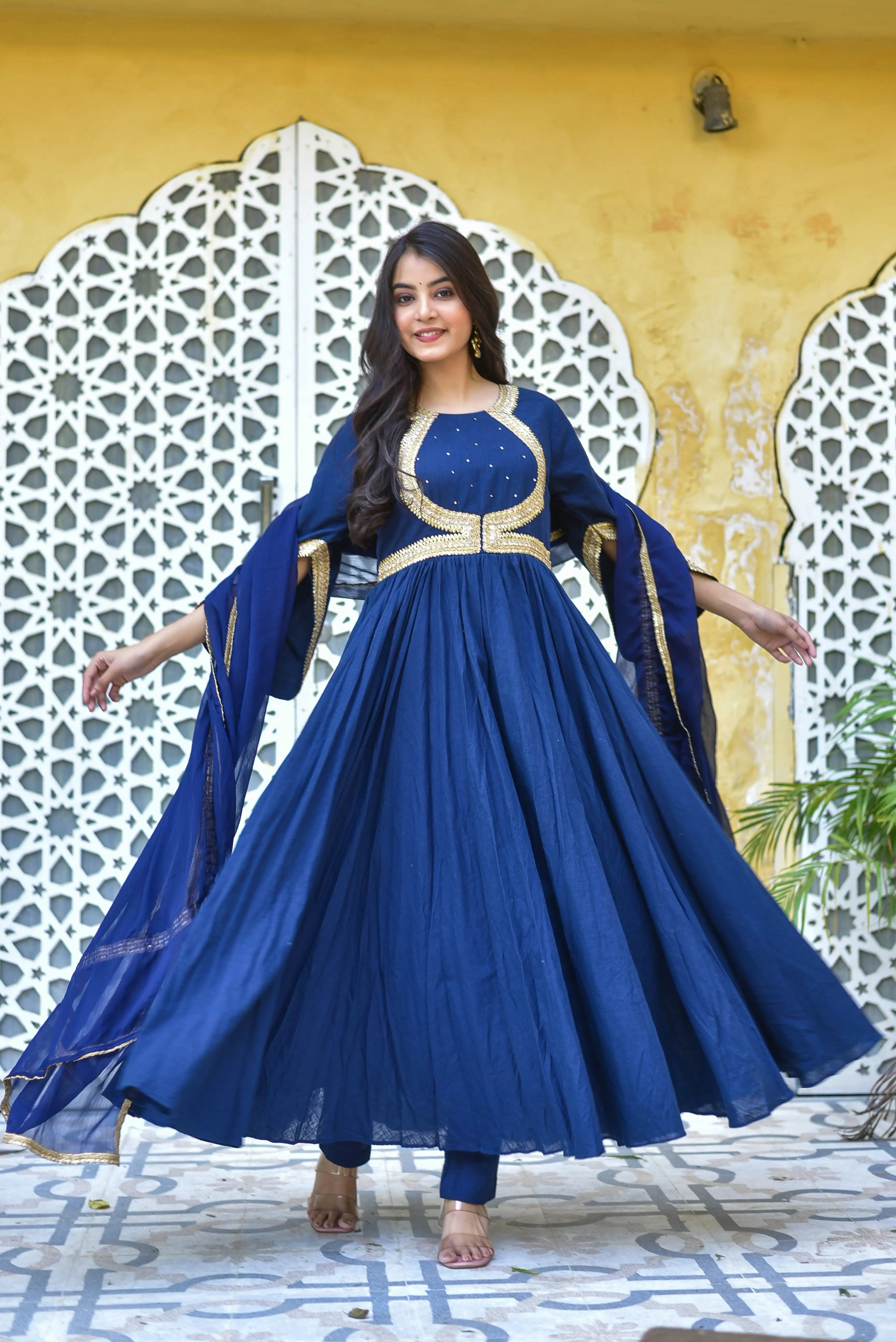 The image depicts a woman wearing a stunning blue anarkali dress, which is a traditional Indian garment. The dress features a round neckline and long sleeves with flared cuffs, and is made of a lightweight fabric that appears to be silk or chiffon. The dress is adorned with intricate gold embroidery, known as Mukesh work, which adds an extra layer of elegance to the overall design.
The dress is a deep blue color, which complements the woman's skin tone and adds a touch of sophistication
