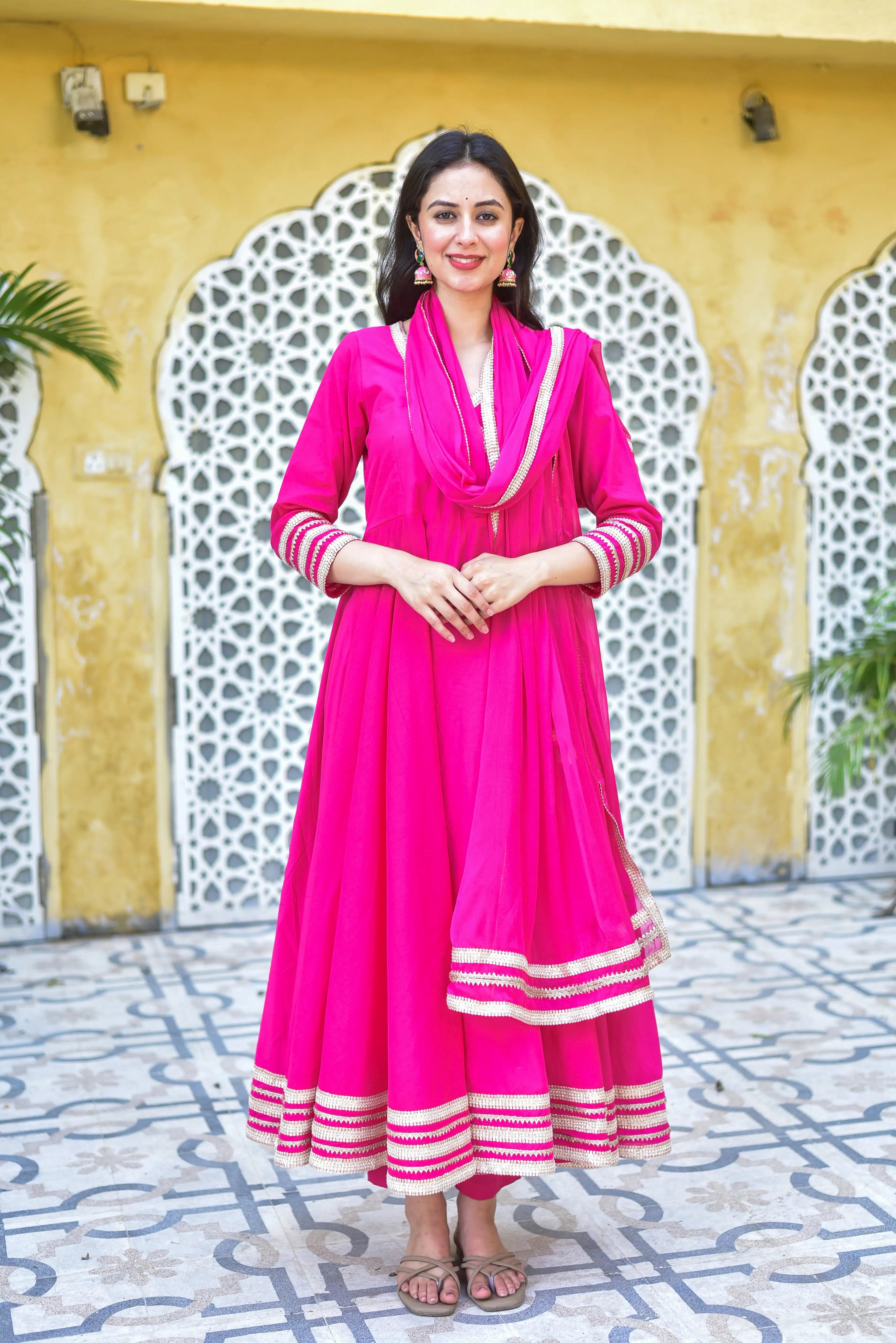The image showcases a stunning rani pink anarkali set, exuding vibrant elegance and regal charm. The rich fuchsia hue of the outfit is accentuated by intricate gold trimmings, adding a touch of sophistication to the overall design. The anarkali style, characterized by its flowing silhouette and flared sleeves, creates a dramatic and feminine silhouette that is perfect for making a statement at any occasion.