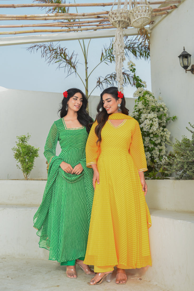 Two women in green and yellow traditional outfits standing on a white staircase with plants in the background.