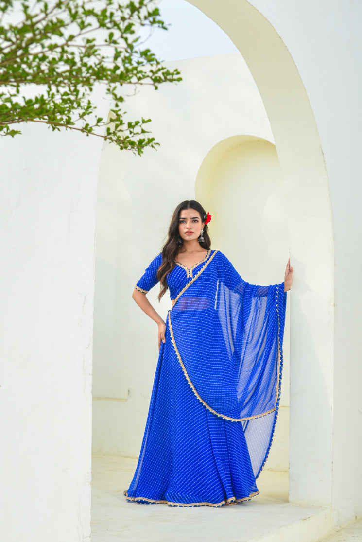 Woman in a blue saree standing in front of a white archway with greenery.