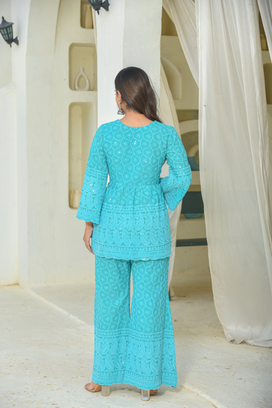 Woman wearing a turquoise embroidered outfit standing in a room with white curtains.