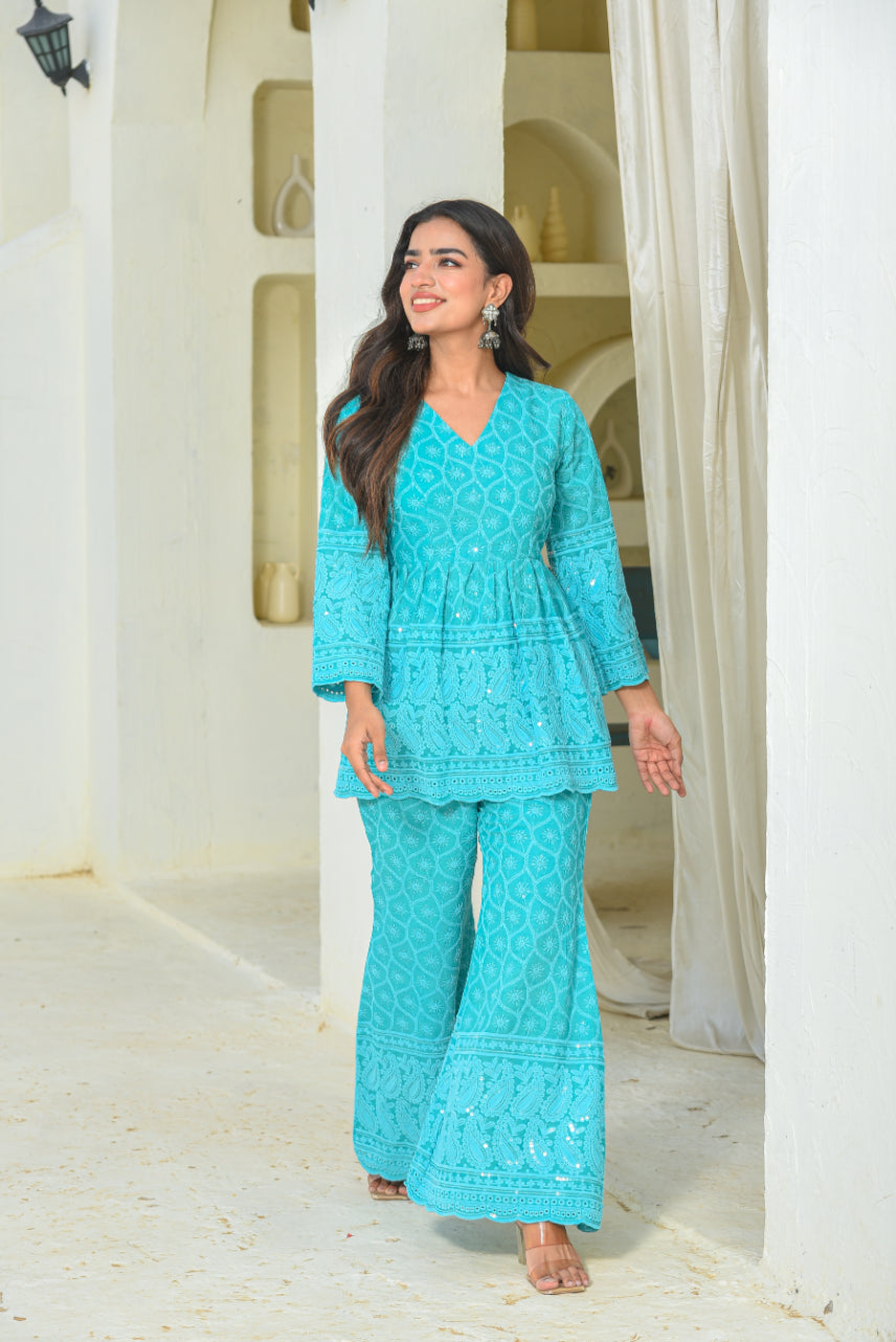 Woman wearing a turquoise traditional outfit standing in a bright, sunlit room with white columns.