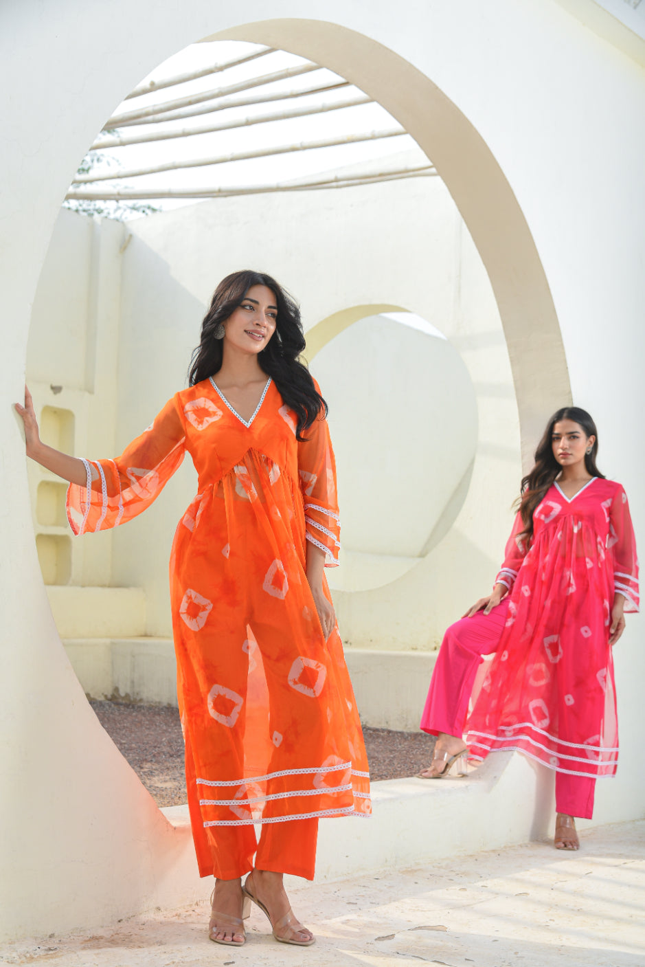 Two women in orange and pink traditional outfits standing in front of a white architectural background.