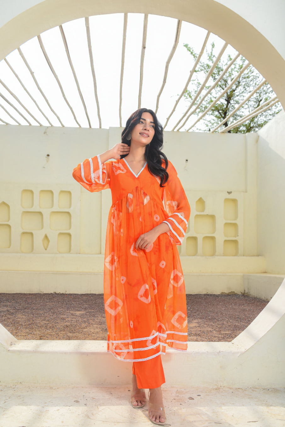 Woman in an orange dress standing in a circular architectural structure with a white wall and tree in the background.