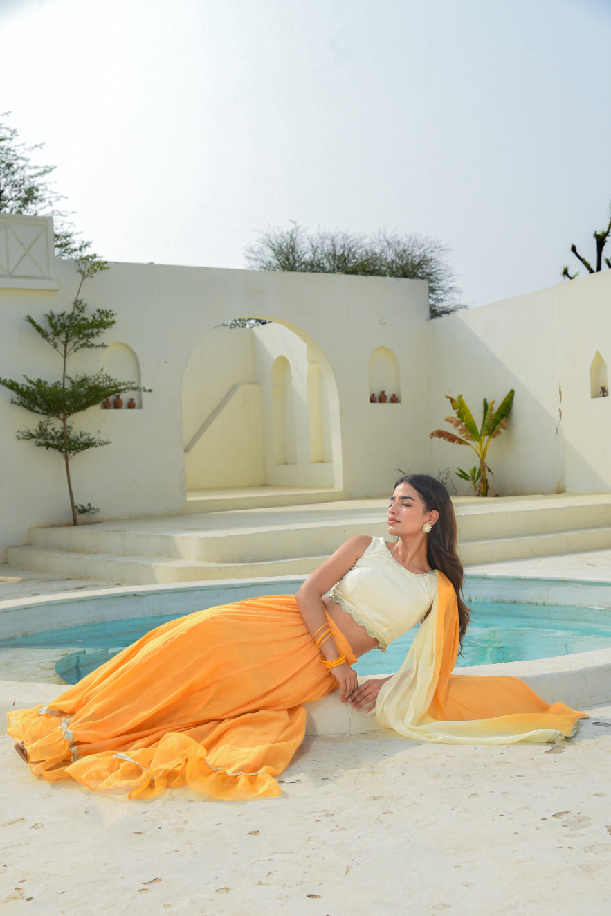 Woman in a white and orange saree sitting by a pool with a white architectural background