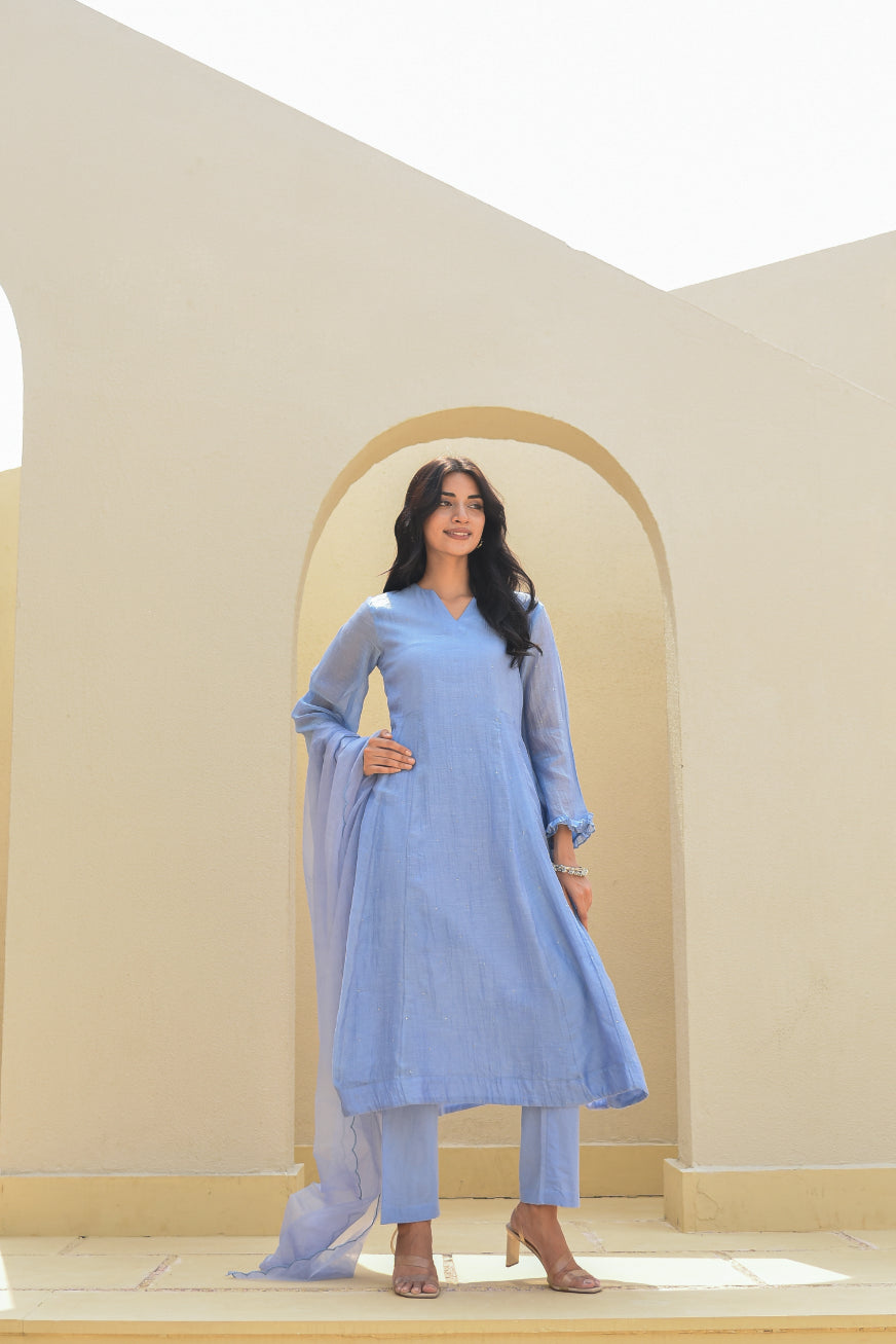 Woman in a light blue traditional outfit standing in a neutral-colored room with an archway.