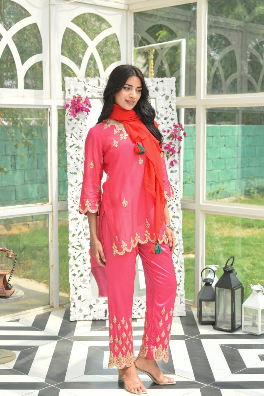 Woman in a pink traditional outfit with a red dupatta standing in a decorative indoor setting.