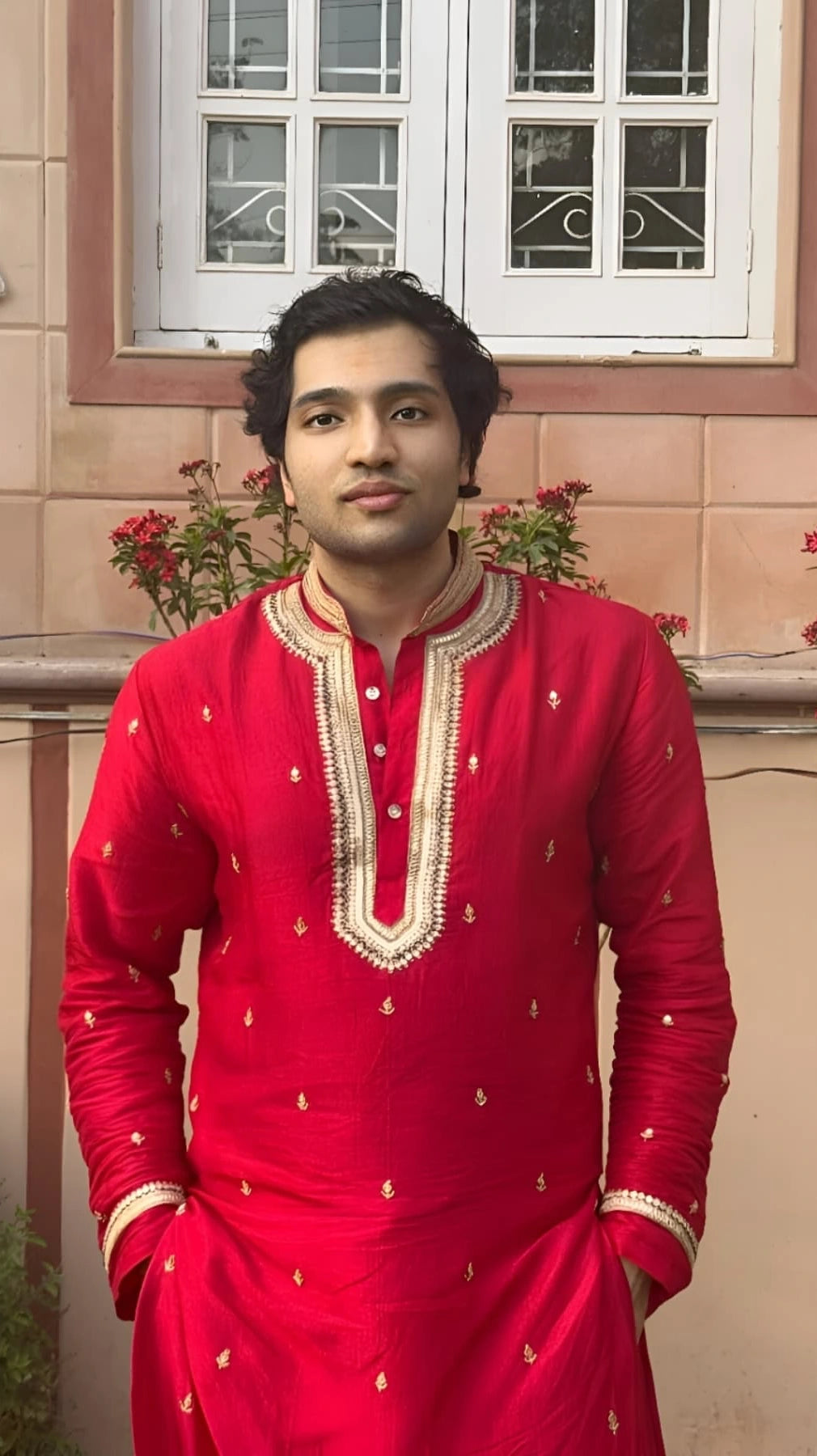 Man wearing a red traditional outfit standing in front of a window with decorative elements.