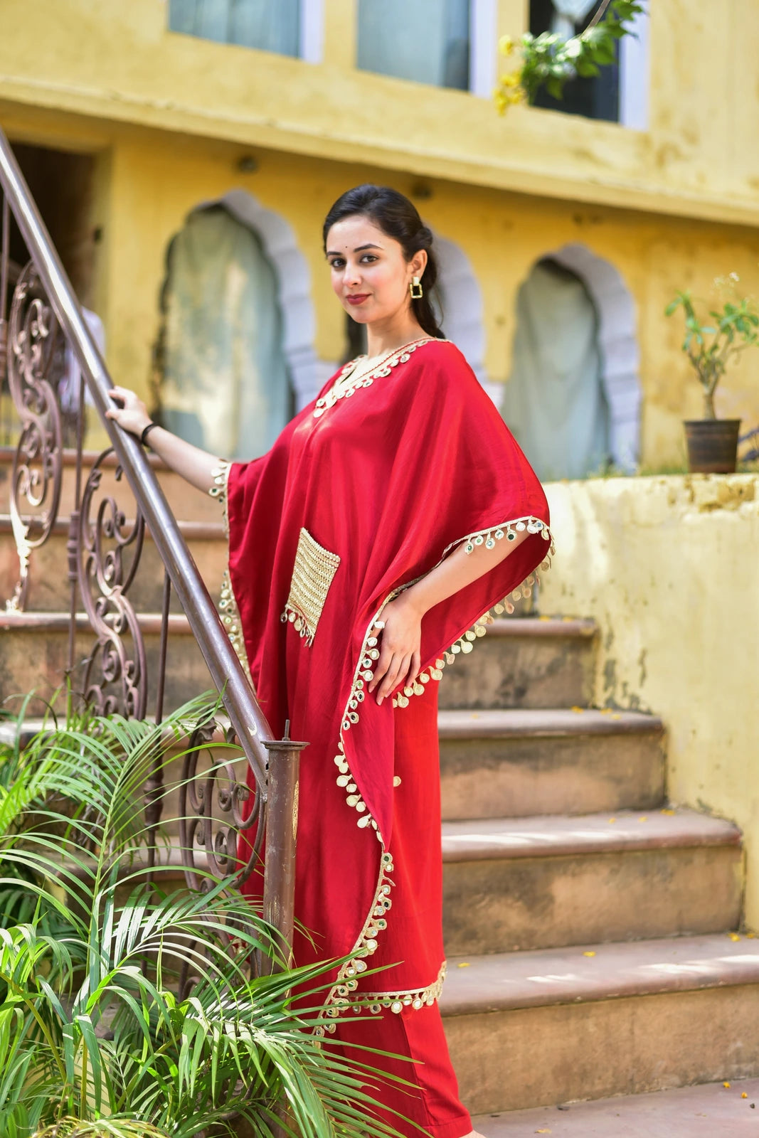 The image showcases a woman in a striking red kaftan, a traditional Middle Eastern garment, standing on a staircase in front of a yellow building. The kaftan features a V-neckline and a loose, flowing design with wide sleeves. It is adorned with intricate gold embroidery along the edges and hem, adding a touch of elegance to the overall look. The woman's dark hair is pulled back, and she accessorizes with a bindi and earrings, complementing her outfit.
The red kaftan is a vibrant and eye-catching piece, pe
