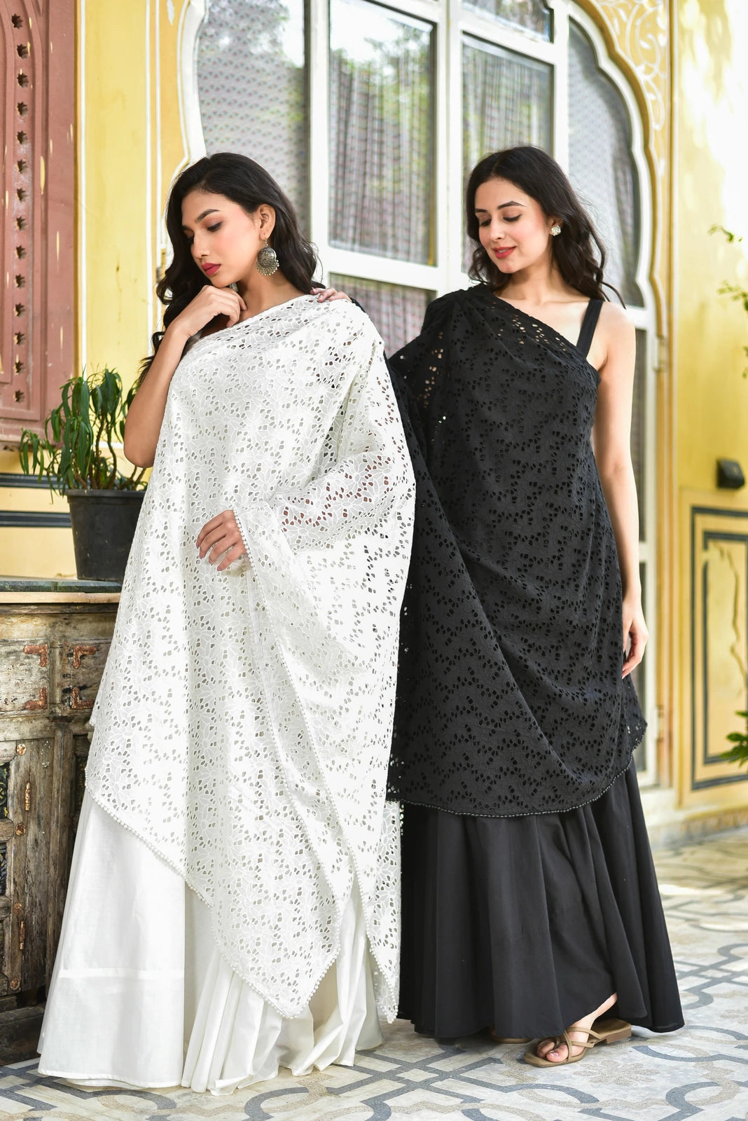 The image showcases two women modeling traditional Indian attire, specifically a long skirt and a long, one-shoulder tunic with intricate embroidery.
The woman on the left wears a white outfit featuring a long, white skirt paired with a white embroidered tunic. The tunic boasts an intricate pattern of small, circular cutouts, which is characteristic of the "Mukesh work" technique. This style of embroidery is known for its delicate and ornate design.
In contrast, the woman on the right
