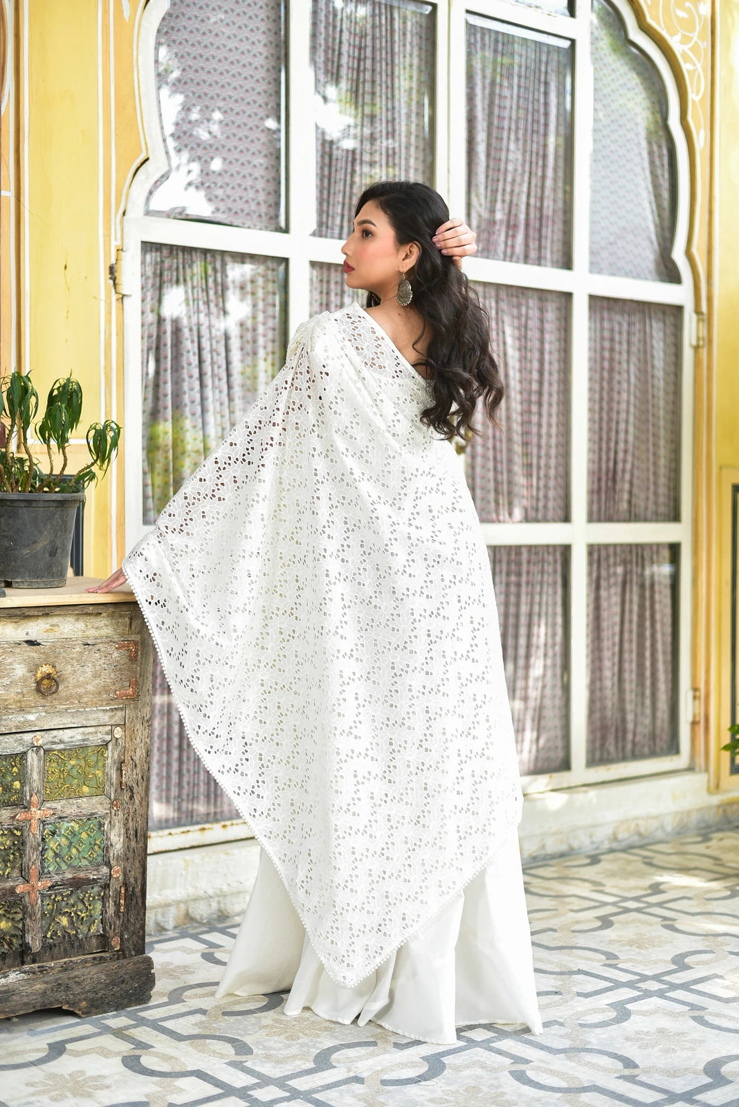 The image depicts a woman standing in front of a window, wearing a white dress and cape. The woman has long dark hair and is facing to the left, with her right hand on her head and her left hand on a wooden cabinet. She is wearing a white dress with a lace cape over it, and large silver earrings.
The background of the image is a yellow wall with a large window featuring white trim and curtains. The floor is made of white and gray tiles with a geometric pattern.