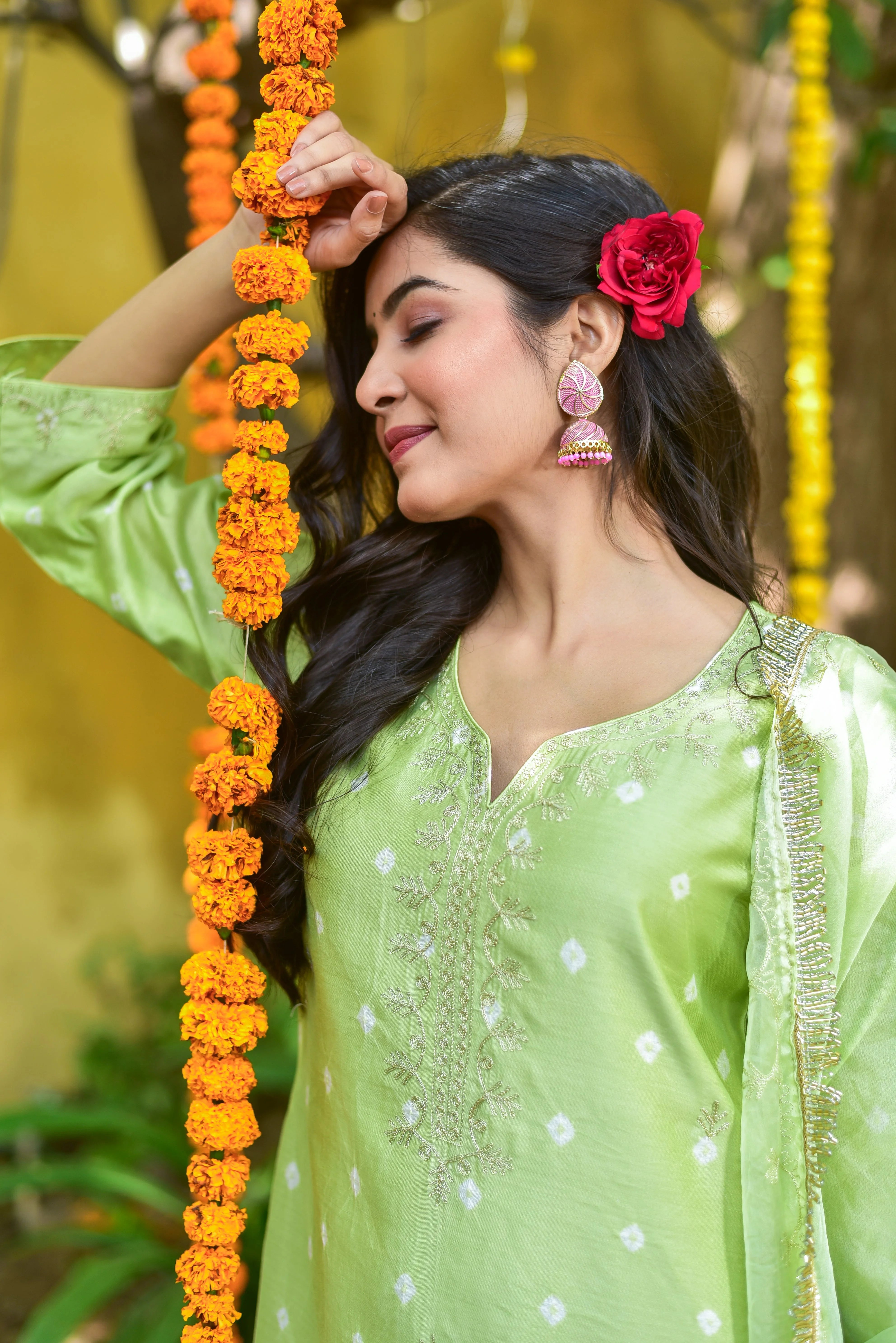 This is a product image for an e-commerce platform, showcasing a woman wearing a green bandhej a-line kurta set. The image features a woman with long, dark hair and a red flower in her hair, posing in front of a blurred background that appears to be a garden or outdoor setting.

Key Features:

 Color: The woman's attire is a vibrant green color, which adds a pop of color to the overall image.