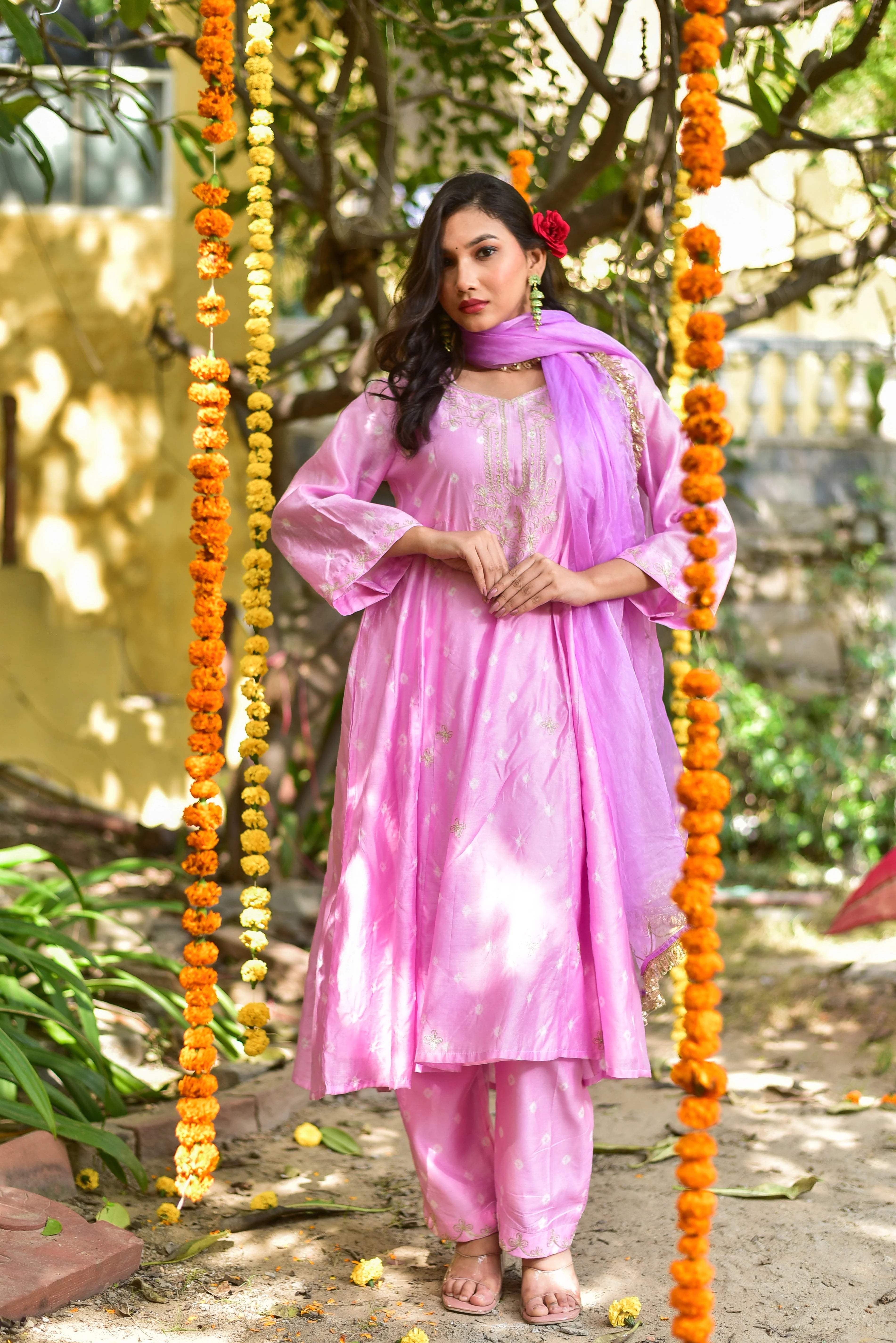 The image depicts a woman wearing a pink salwar kameez, a traditional Indian outfit consisting of a long tunic (kameez), loose-fitting pants (salwar), and a matching dupatta (scarf). The kameez features a round neckline, long sleeves, and a straight cut, while the salwar is loose-fitting and straight-legged. The dupatta is draped elegantly over her shoulders.

The outfit is adorned with intricate Mukesh work, a type of embroidery