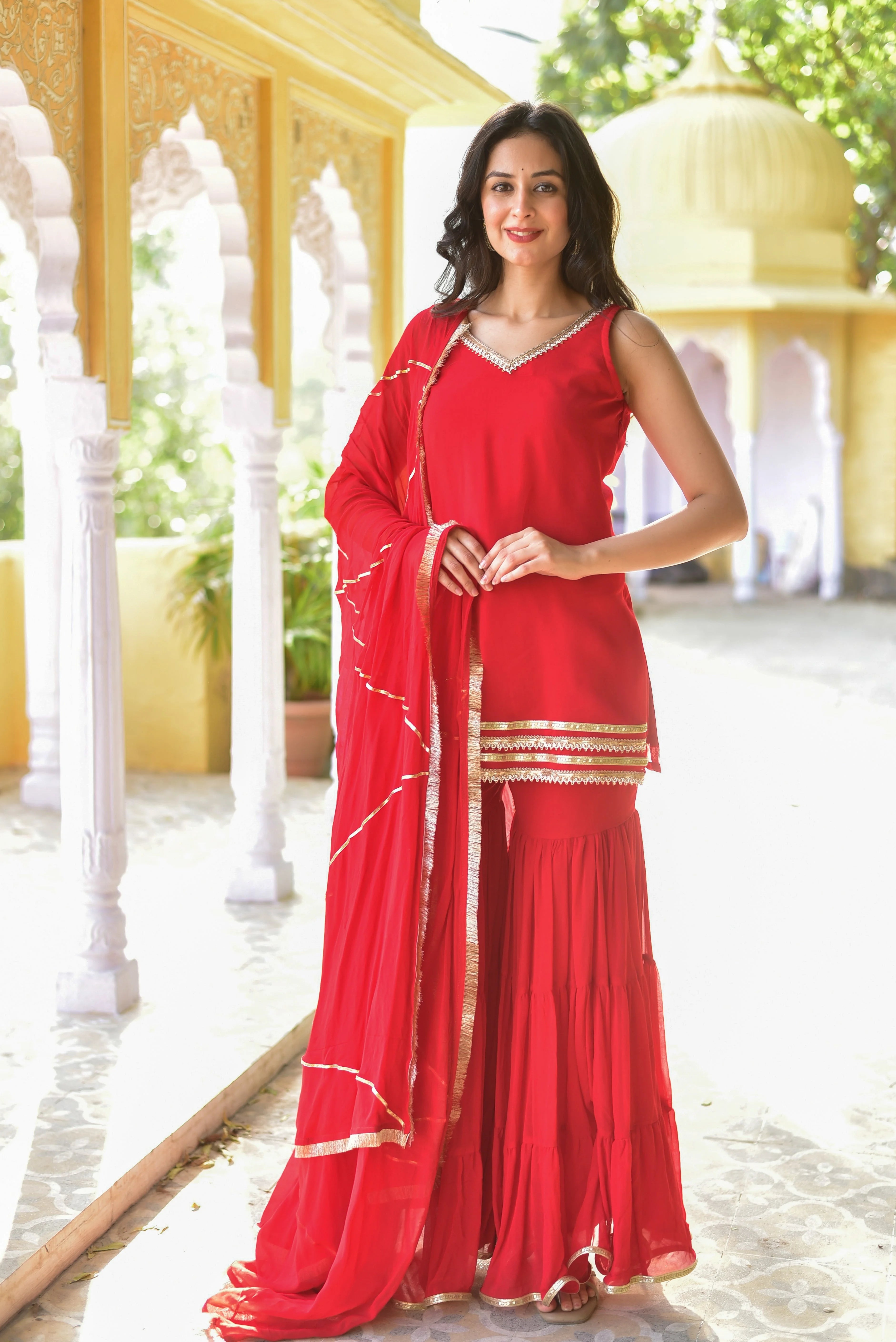 Woman in a red saree standing on a sunlit patio with white pillars and greenery.