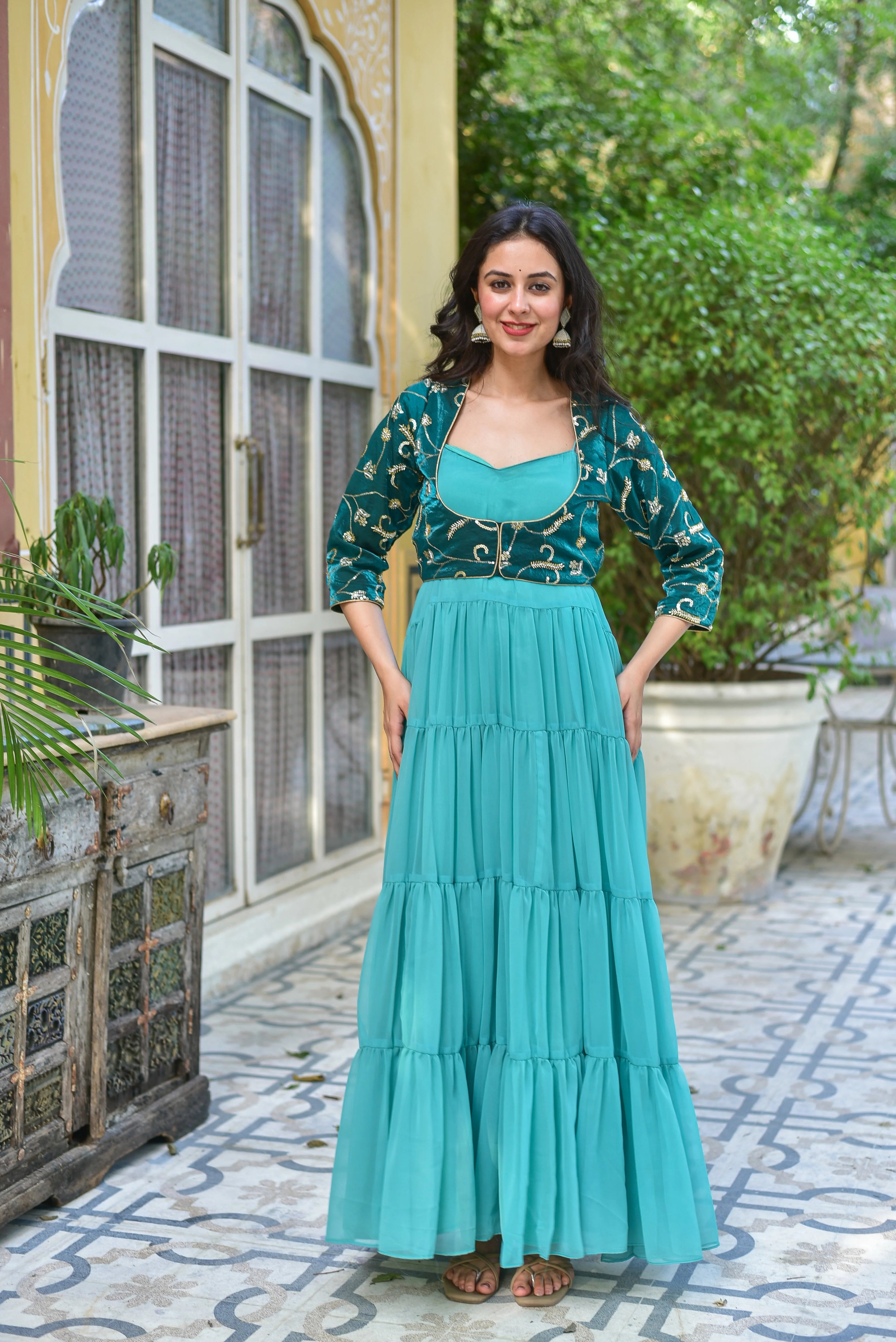 The product is a stunning green tiered dress with a jacket, featuring a contemporary silhouette and traditional craftsmanship. The flowing georgette dress boasts graceful tiers that create beautiful movement with every step, while the coordinating jacket adds a touch of elegance to the overall ensemble. This exquisite outfit is perfect for making a statement at any celebration, combining modern style with timeless charm.