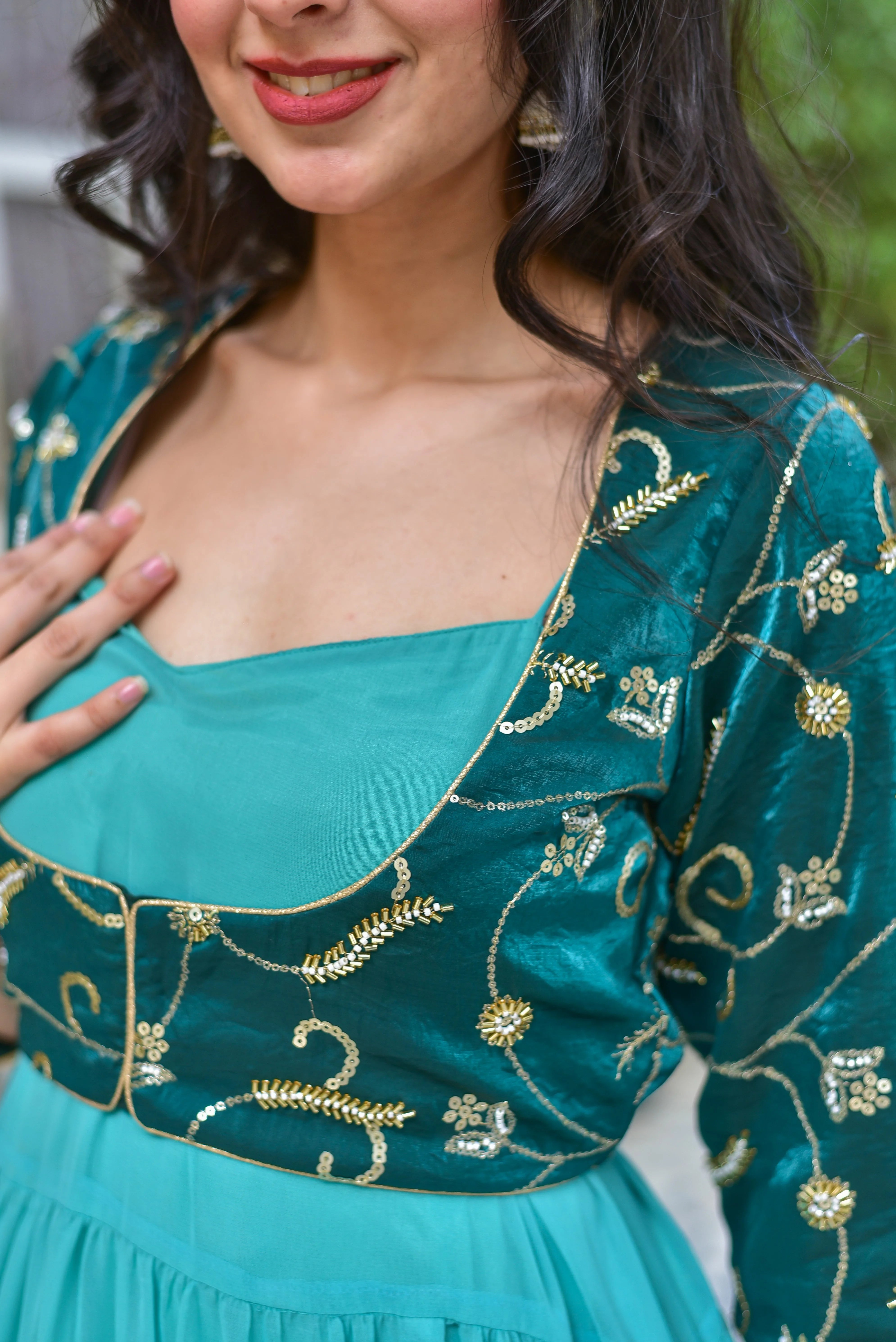 The image showcases a stunning green dress with a jacket, featuring a contemporary silhouette and traditional craftsmanship. The flowing georgette dress boasts graceful tiers that create beautiful movement with every step, while the coordinating jacket adds an extra touch of elegance. The dress is adorned with intricate gold embroidery, adding a luxurious touch to the overall design. The model's pose and smile convey confidence and poise, making this dress perfect for any special occasion.