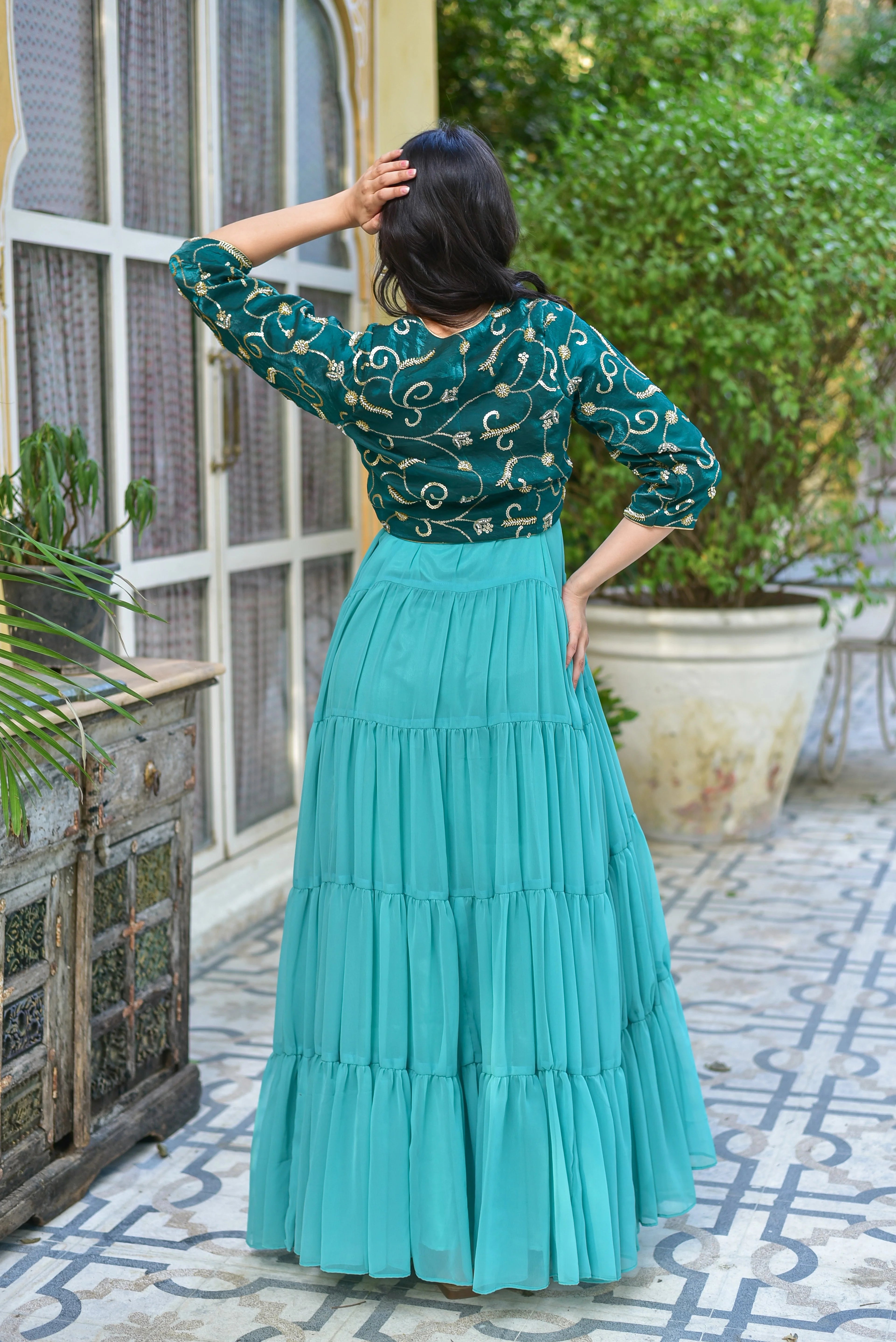 The image shows a woman in a stunning green dress with a jacket, posing in front of a window and plants. The dress is made of a lightweight material and has a tiered design, with each tier slightly shorter than the last. The jacket is a matching green color and has a more fitted silhouette than the dress.

The woman's hair is dark and styled in loose waves, and she is wearing minimal makeup.