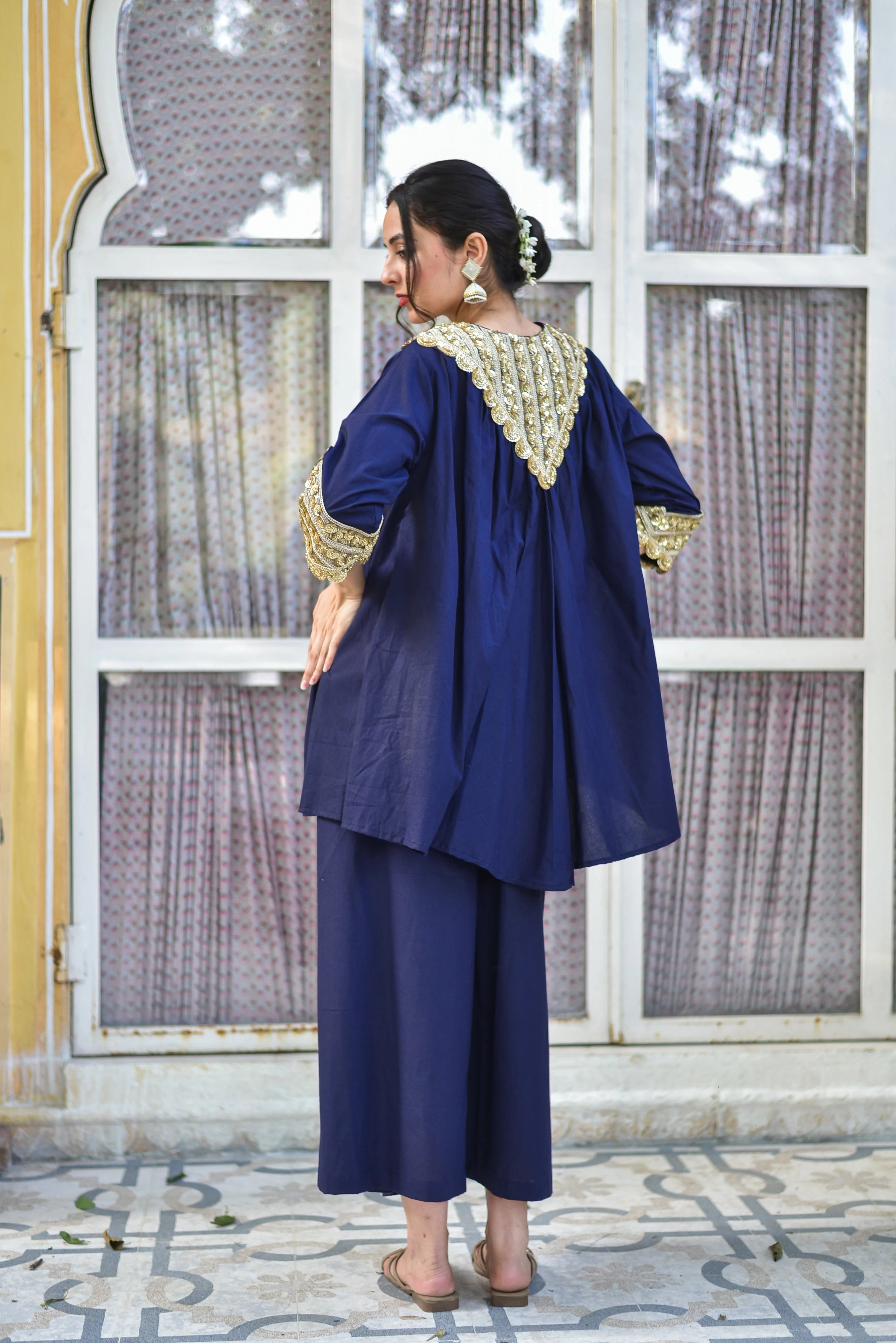 The image showcases a woman modeling a stunning navy blue outfit, exuding elegance and sophistication. The ensemble consists of a long tunic with intricate gold embroidery adorning the collar and sleeves, complemented by a matching pair of wide-legged pants. The tunic features a loose-fitting design with a flowing silhouette, while the pants are cropped just above the ankle. The woman's hair is styled in a low bun, adorned with a delicate white flower, and she accessorizes with large gold earrings. Her
