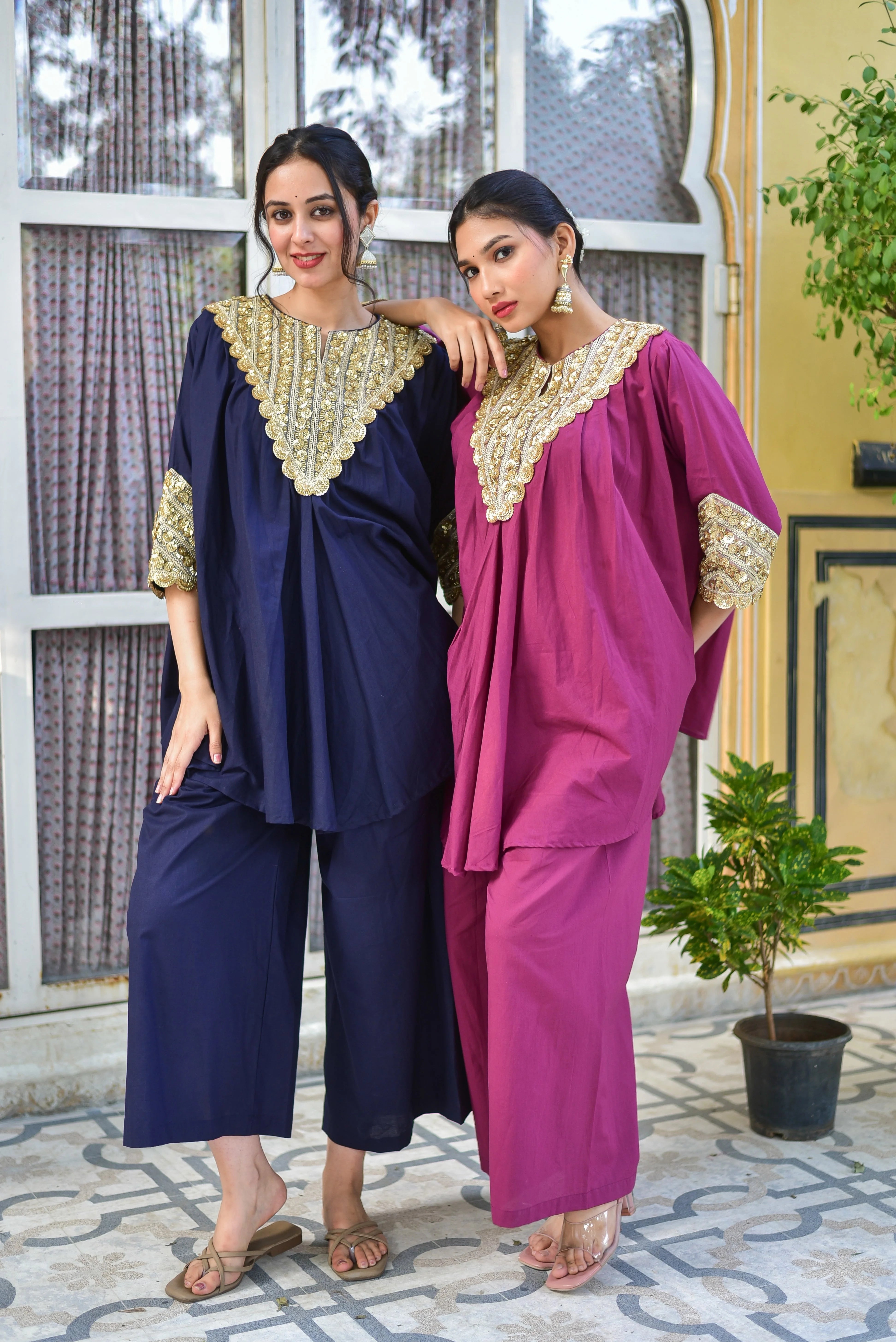 The image showcases two women modeling elegant, traditional Indian attire. The woman on the left wears a navy blue ensemble with gold embroidery and embellishments on the neckline and sleeves, complemented by matching navy blue pants and tan sandals. Her counterpart on the right sports a vibrant pink outfit with matching gold accents, paired with pink pants and transparent sandals. Both outfits feature intricate gold embroidery on the neckline and sleeves, adding to their luxurious appeal. The women's dark 