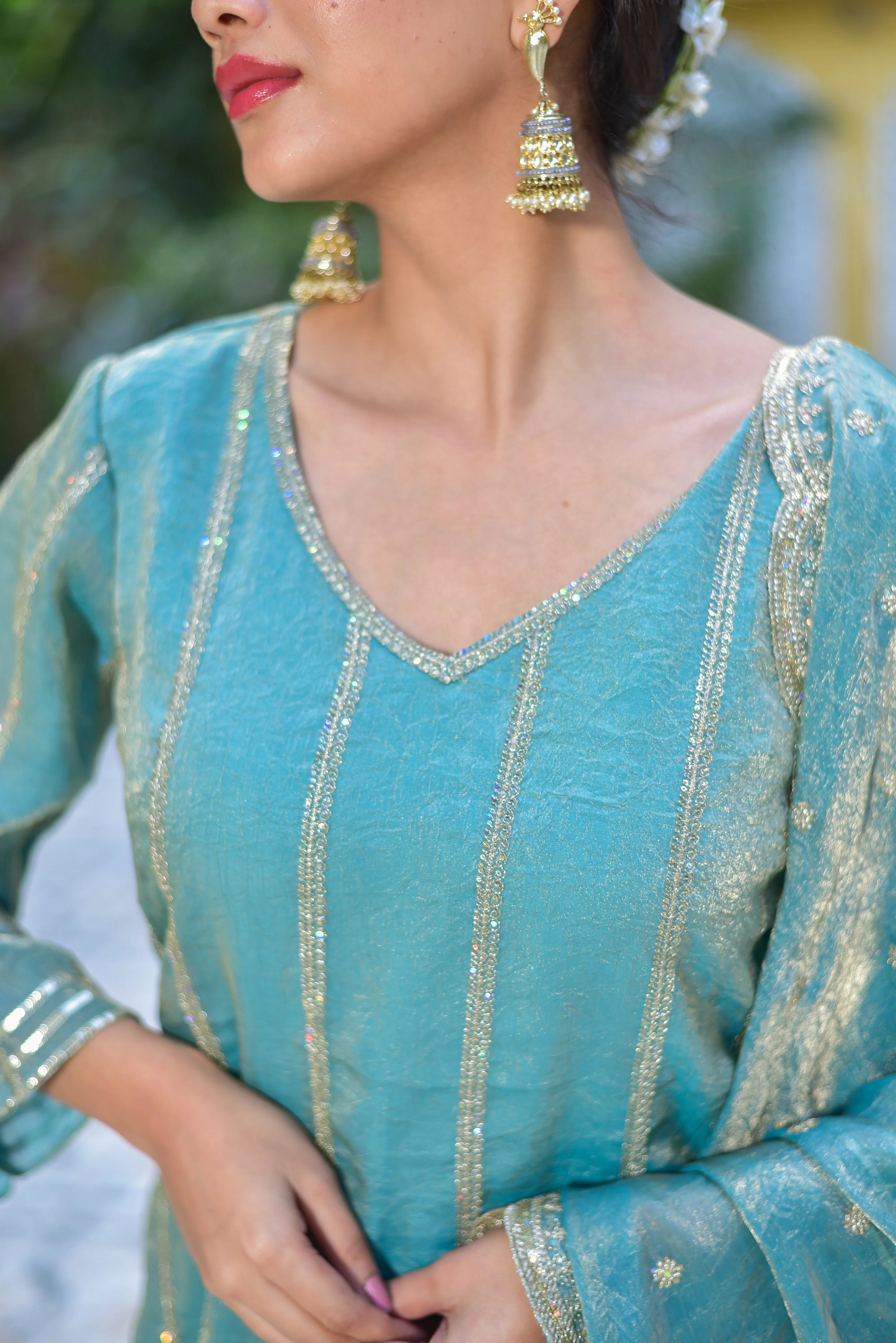 This image showcases a stunning Sea Green Kurta Sharara Set, exuding elegance and sophistication. The sea green color is a beautiful shade, reminiscent of the ocean's tranquil hue. The fabric appears to be a luxurious silk or organza, with a subtle sheen that catches the light.

The kurta features a V-neckline, adorned with intricate gold embroidery that adds a touch of regality to the overall design. The sleeves are elegantly flared, with a slight bell shape at the end, creating a dramatic effect.