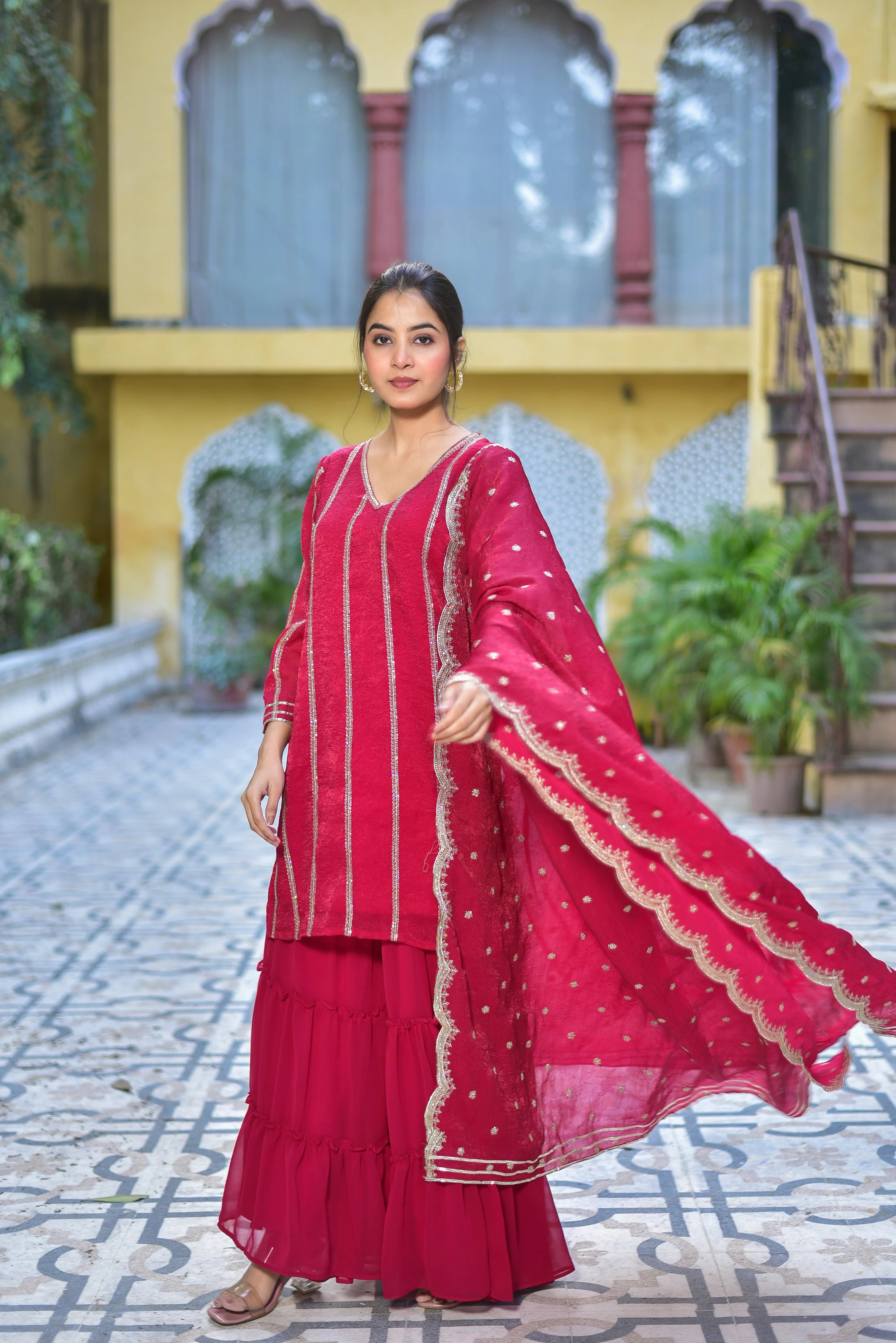 The image showcases a woman modeling a vibrant Magenta Heavy Sharara Set, exuding elegance and sophistication. The set features a long, red kurta with silver embroidery, complemented by a matching sharara and a red dupatta adorned with intricate gold embroidery. The woman's dark hair is pulled back, and she wears gold earrings, adding a touch of glamour to the overall look.