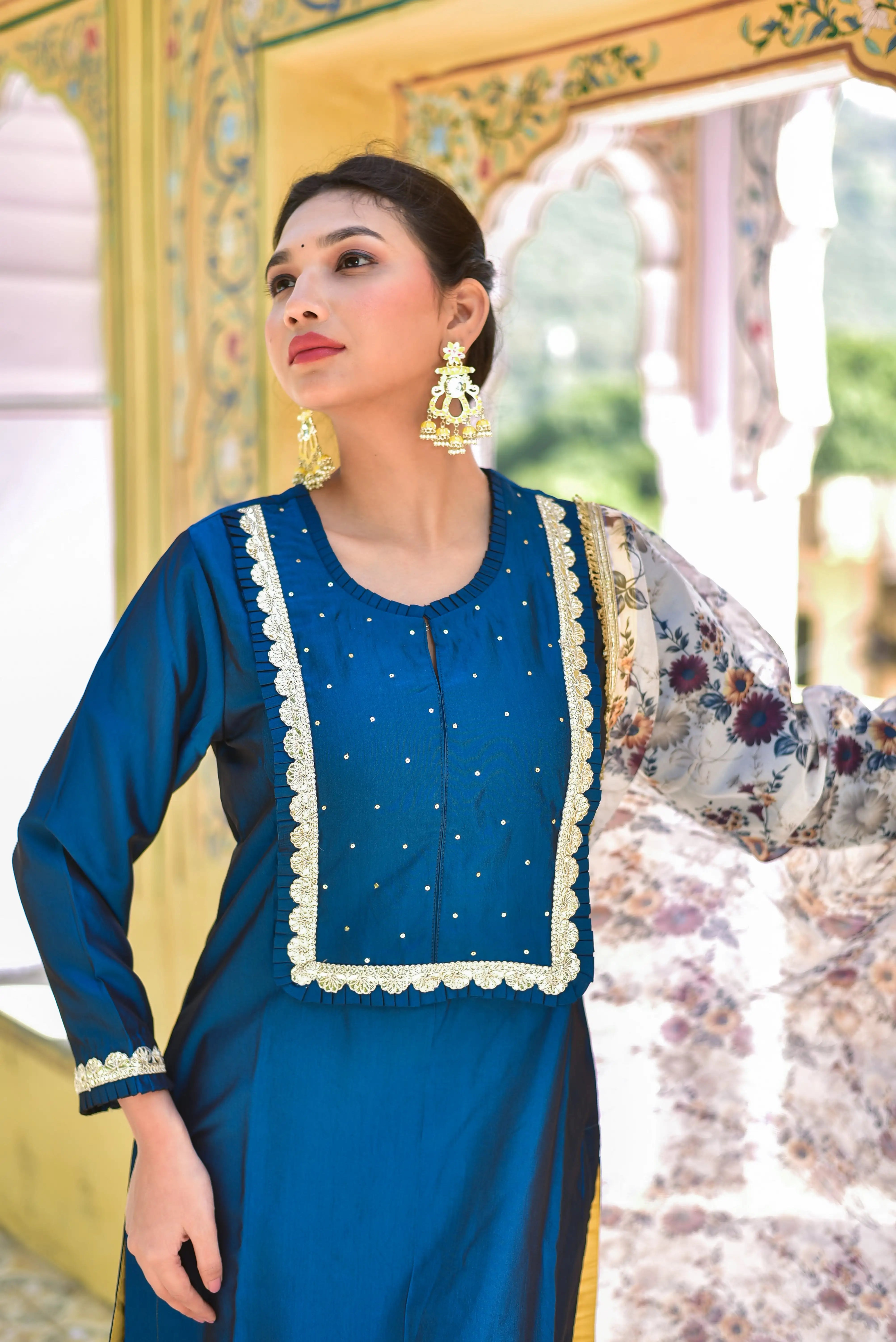 The image showcases a woman adorned in a stunning peacock blue kurta and pant set, complemented by a beautiful floral dupatta. The kurta features intricate gold embroidery on the neckline and cuffs, adding a touch of elegance to the overall design. The woman's dark hair is styled in a neat bun, and she wears elegant gold earrings that add a pop of sophistication to her overall look.