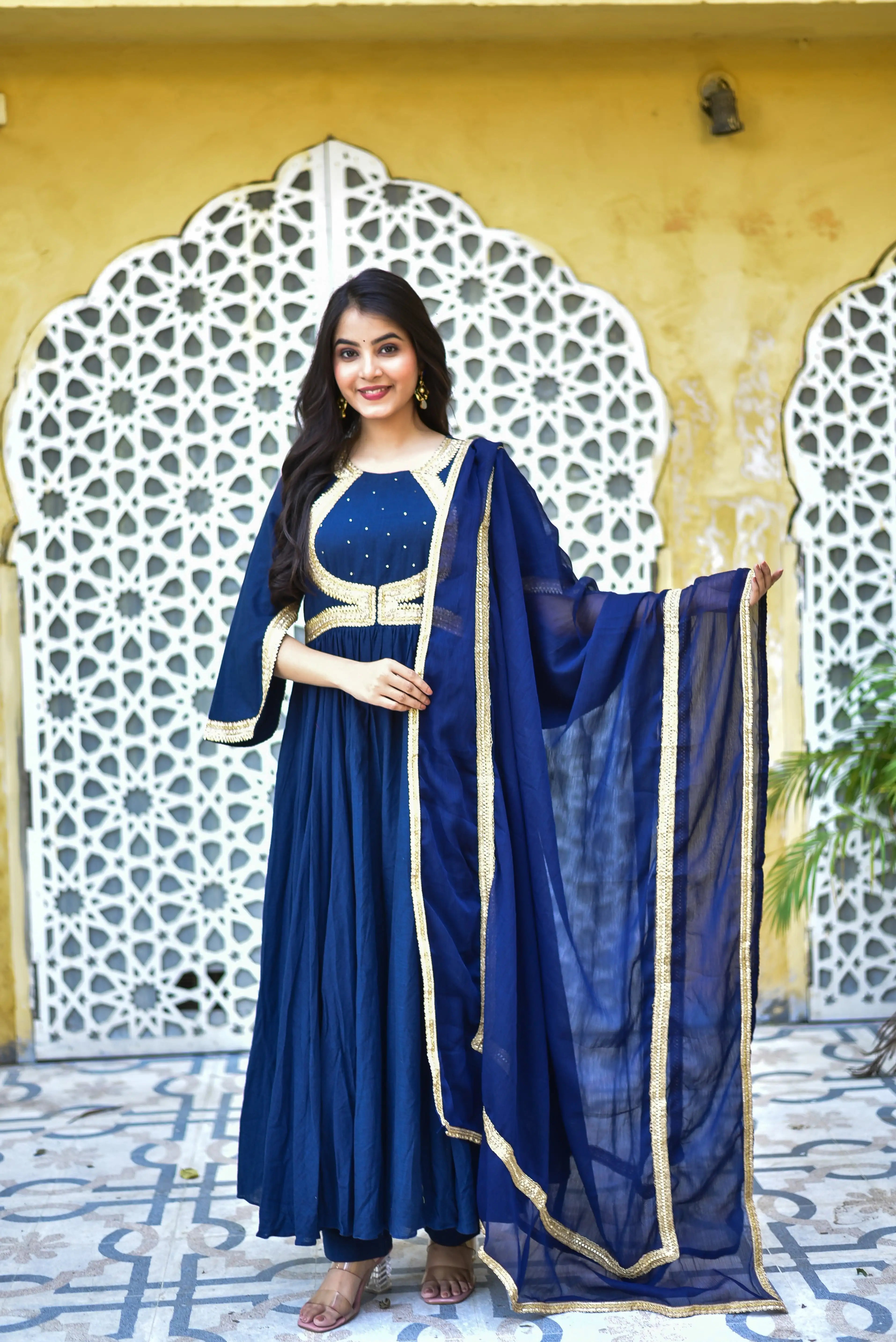 The image showcases a woman modeling an elegant blue Anarkali dress, adorned with intricate gold embroidery and a matching dupatta.

The Anarkali dress is a floor-length, long-sleeved, and flowing garment that features a round neckline and a gold-embroidered bodice. The dress is crafted from a lightweight fabric, likely silk or chiffon, and boasts a subtle sheen. The gold embroidery adds a touch of opulence, with delicate patterns and designs that catch the light.