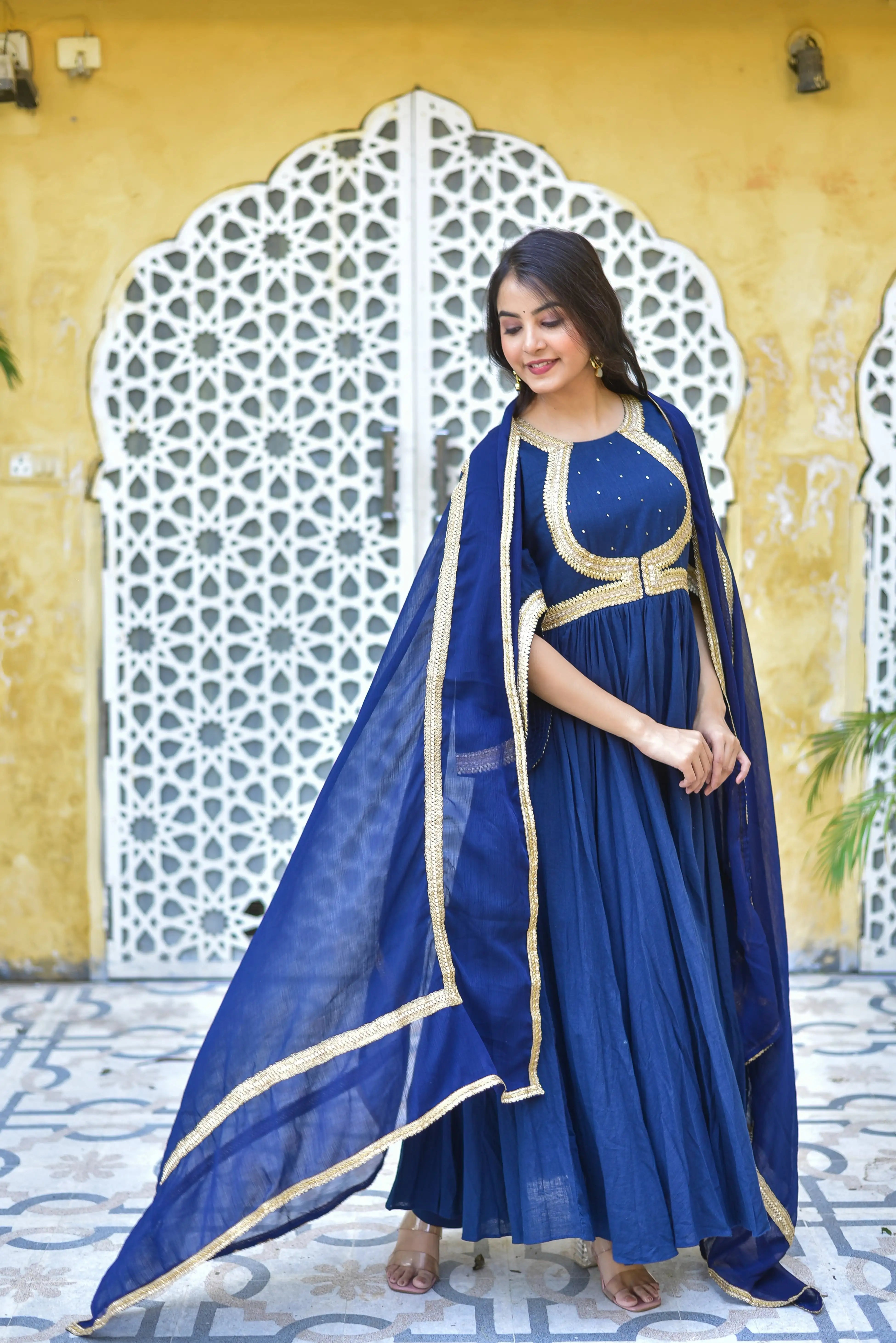 The image features a woman wearing a stunning blue dress with gold accents, exuding elegance and sophistication. The dress is a floor-length, ankle-length design, crafted from a lightweight fabric such as cotton or silk, and features a round neckline and three-quarter sleeves. The bodice is adorned with intricate gold embroidery, including Mukesh work, which adds a touch of luxury and opulence.

The dress is paired with a matching blue dupatta, also featuring gold trim, which adds a pop of color and