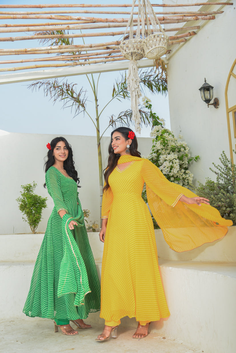 Two women in green and yellow traditional outfits standing outdoors with a decorative arch and plants in the background.