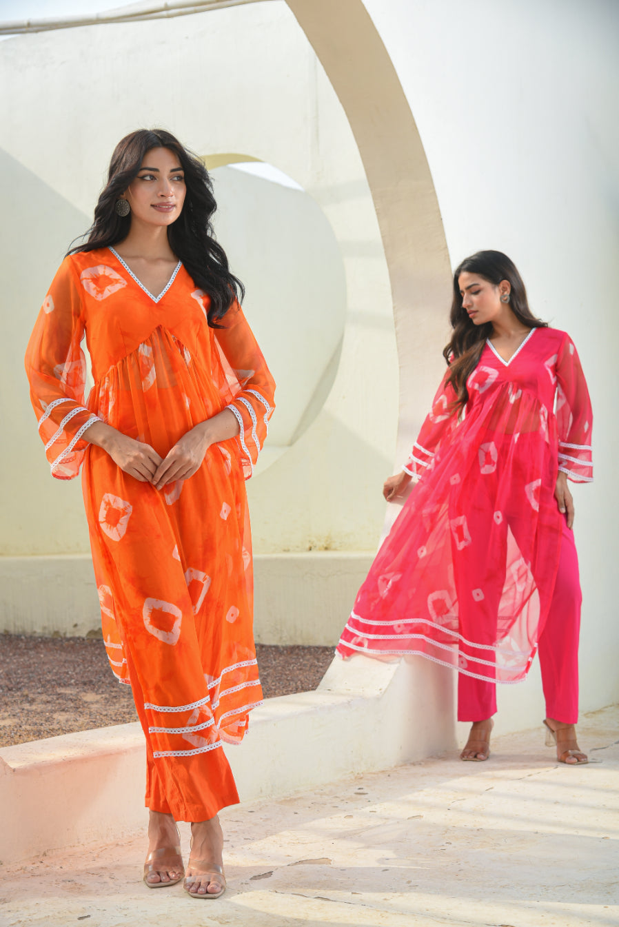 Two women wearing orange and pink outfits with white patterns, standing against a white architectural background.