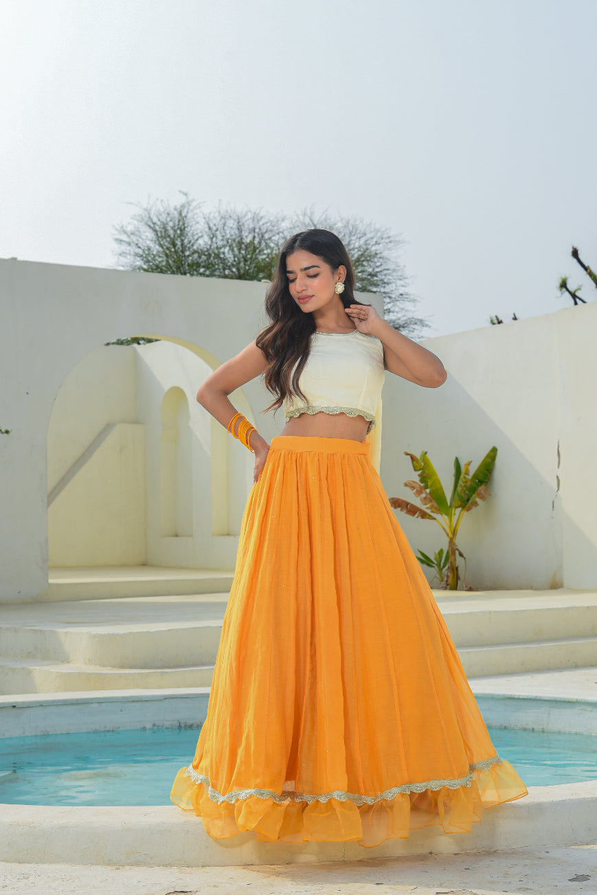 Woman in a white top and orange skirt standing by a pool with a white wall and greenery in the background