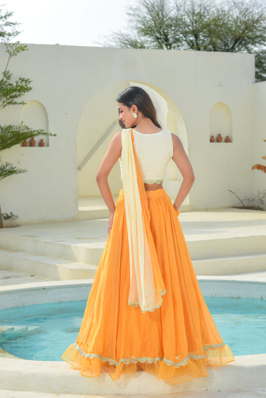Woman in an orange and white traditional outfit standing by a pool with a white building and greenery in the background.