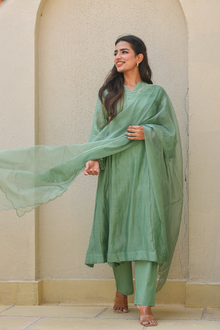 Woman in a green traditional outfit standing against a beige wall.