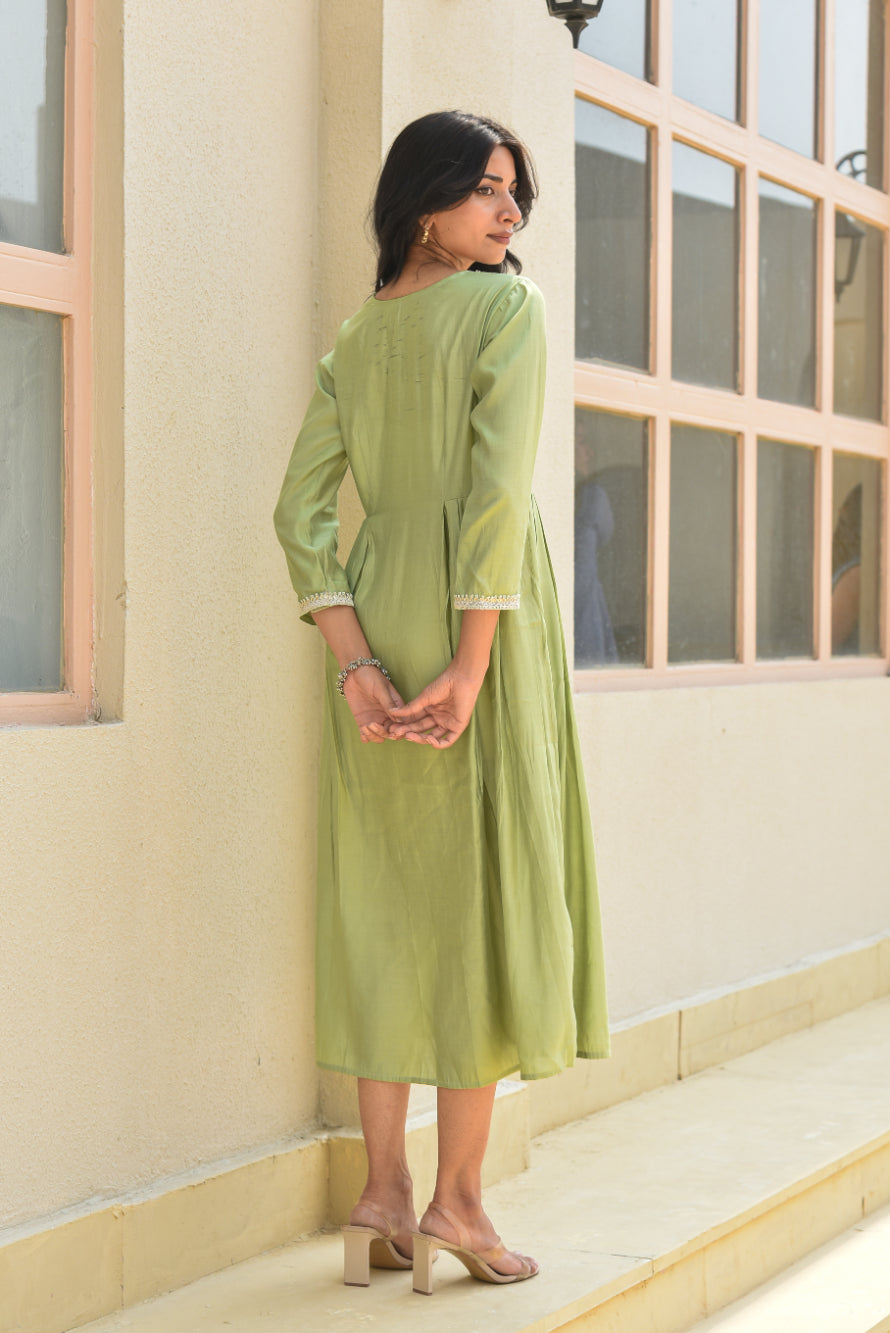 Woman in a green dress standing against a light-colored wall with windows.