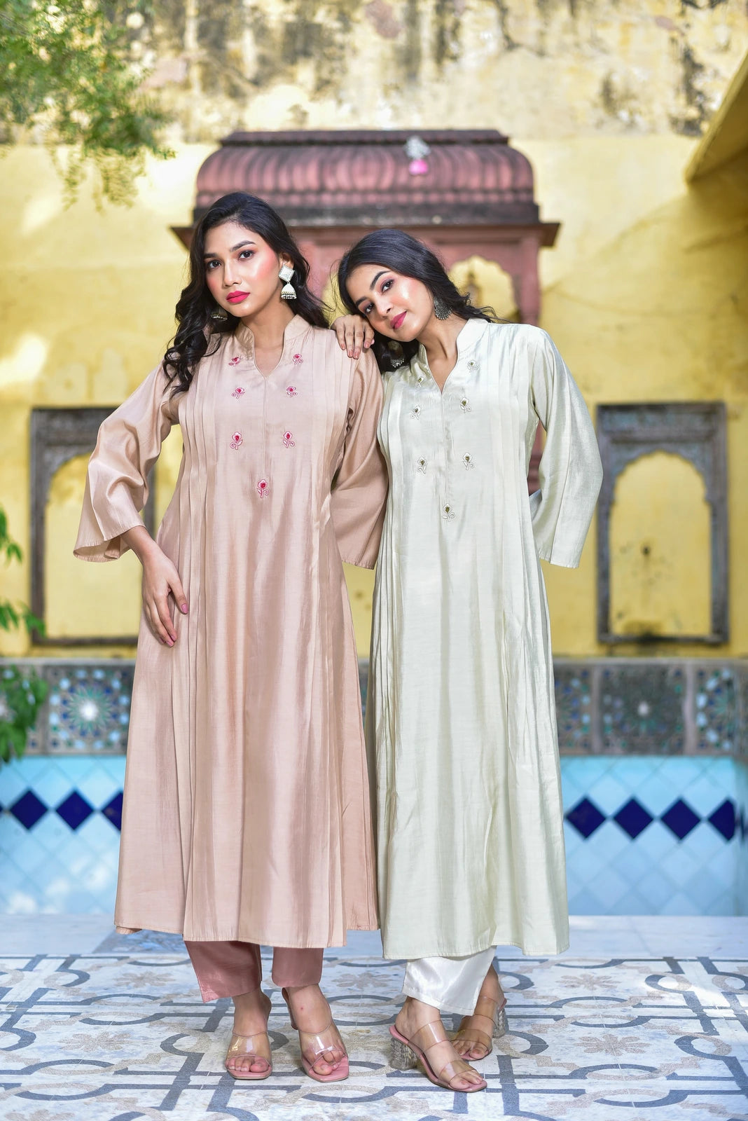 The image features two women posing in a traditional Indian courtyard setting, showcasing the Creamy Kaju Katli Pleated Kurta Set. The woman on the left wears a beige kurta with intricate embroidery and pink pants, while the woman on the right sports a cream-colored kurta with matching pants. Both women have dark hair and are dressed in elegant heels.