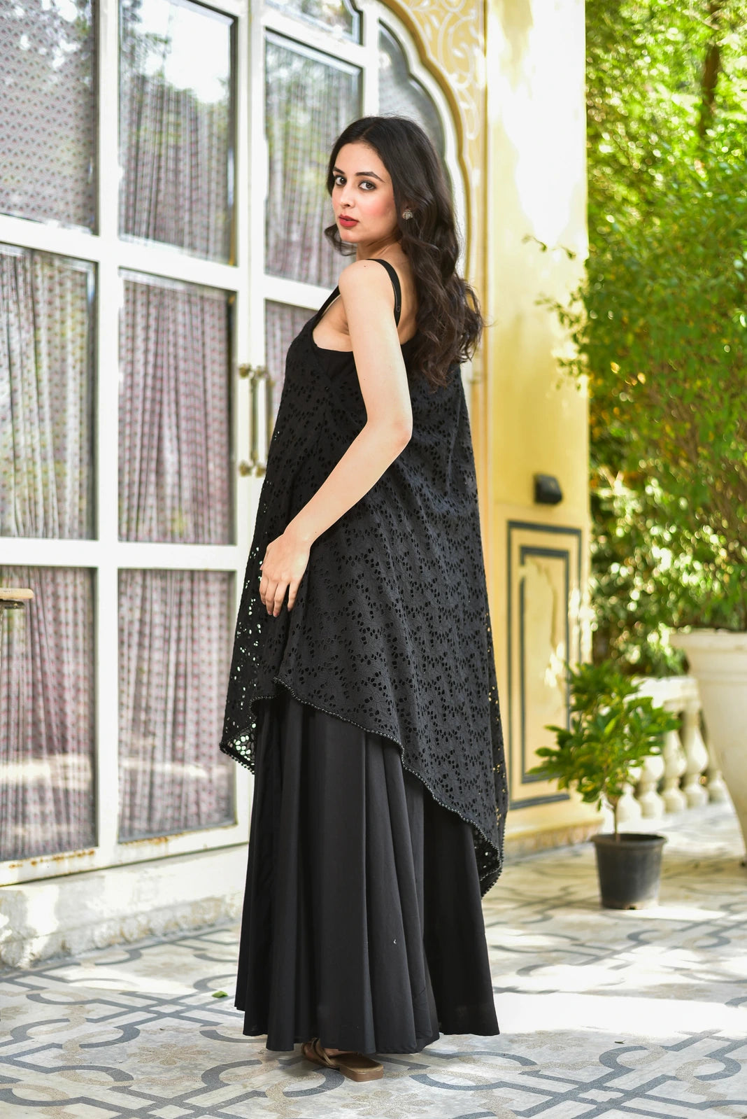The image features a woman showcasing a stunning black dress, exuding elegance and sophistication. The dress boasts a sleeveless design with a high-low hem, crafted from a lightweight fabric that appears to be cotton or silk. A flowing black skirt complements the top, while a long, intricately embroidered tunic-style overlay adds a touch of luxury.

The tunic is adorned with eyelet embroidery, a traditional technique that involves cutting out small circles of fabric to create a delicate pattern. This intric