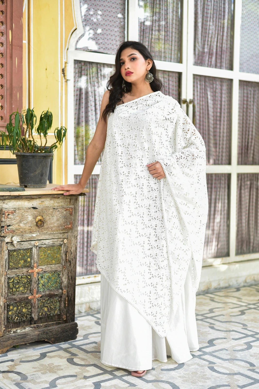 The image features a woman dressed in a stunning white sari, exuding elegance and sophistication. The sari is crafted from a lightweight, semi-sheer fabric, likely cotton or silk, and boasts a delicate eyelet pattern that adds to its charm. The intricate embroidery, known as Mukesh work, is a testament to the artisan's skill and attention to detail.

The woman's attire is completed with a crisp white blouse, which provides a subtle contrast to the sari's intricate design.