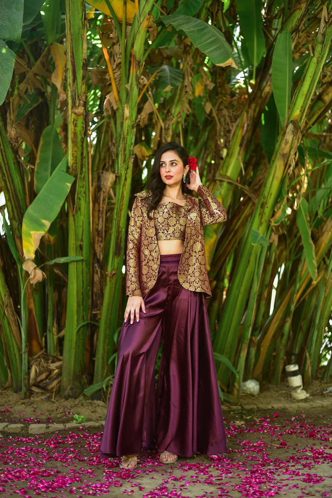 a woman standing in front of a lush backdrop of green plants, exuding an air of elegance and sophistication. She is dressed in a stunning plum-colored three-piece set, comprising a brocade blazer, a matching crop top, and wide-legged pants, all crafted from luxurious fabrics that catch the light beautifully.

The blazer, with its intricate gold embroidery, adds a touch of opulence to the overall ensemble, while the crop top and pants provide a sleek and modern contrast.