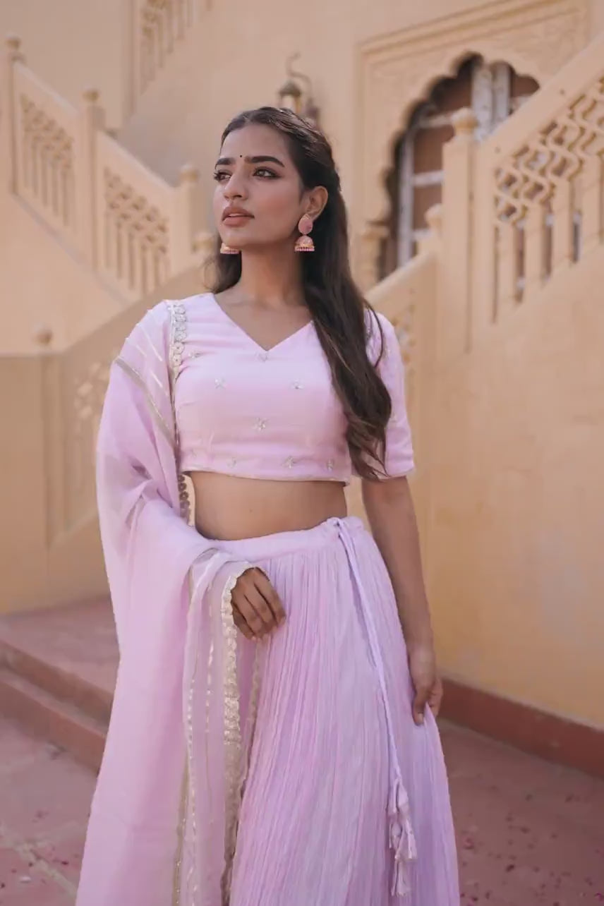 This lilac set features a silk muslin blouse with a V-neckline, half sleeves, and side zip closure, adorned with delicate handwork. It pairs beautifully with a crushed-technique silk organza lehenga with a string closure and a matching organza dupatta—perfect for elegant festive occasions.